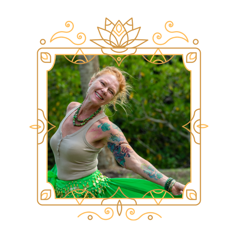 A woman with tattoos and red hair smiling outdoors, wearing a beige top, a green skirt with coins, and a beaded necklace, standing in front of green foliage.