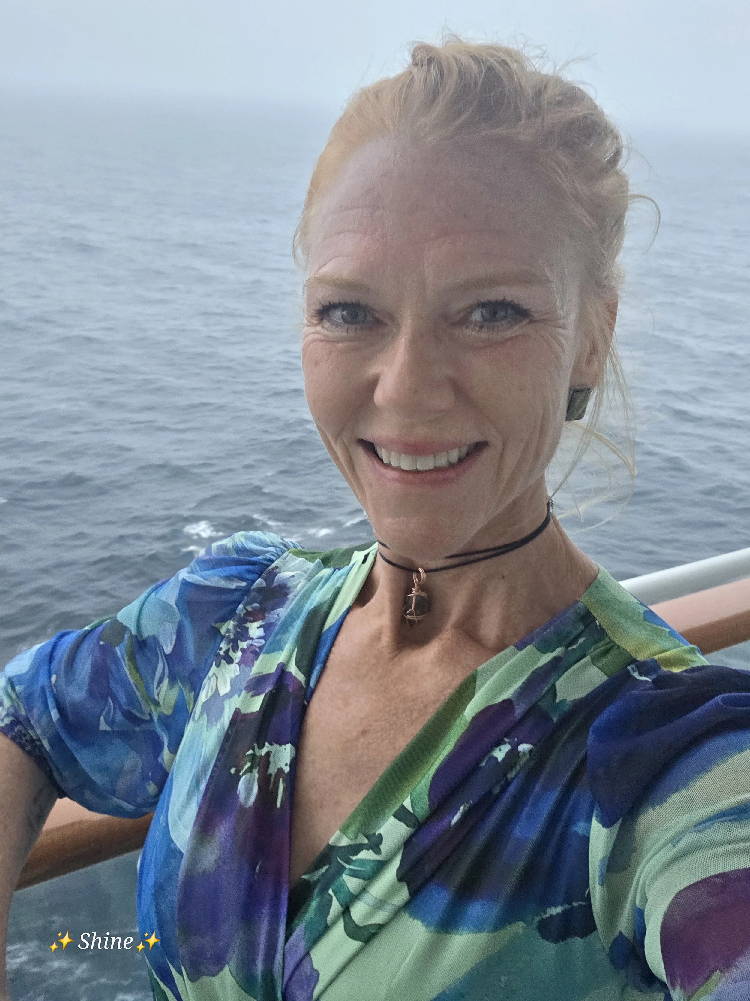 A smiling woman with blue eyes and blonde hair taking a selfie on a boat with the ocean in the background, wearing a colorful patterned top and a choker necklace.