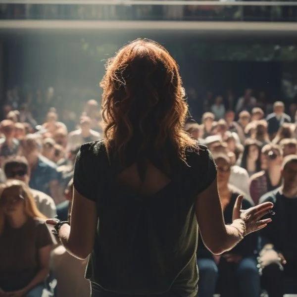 A woman standing on stage facing a large audience at an outdoor event.