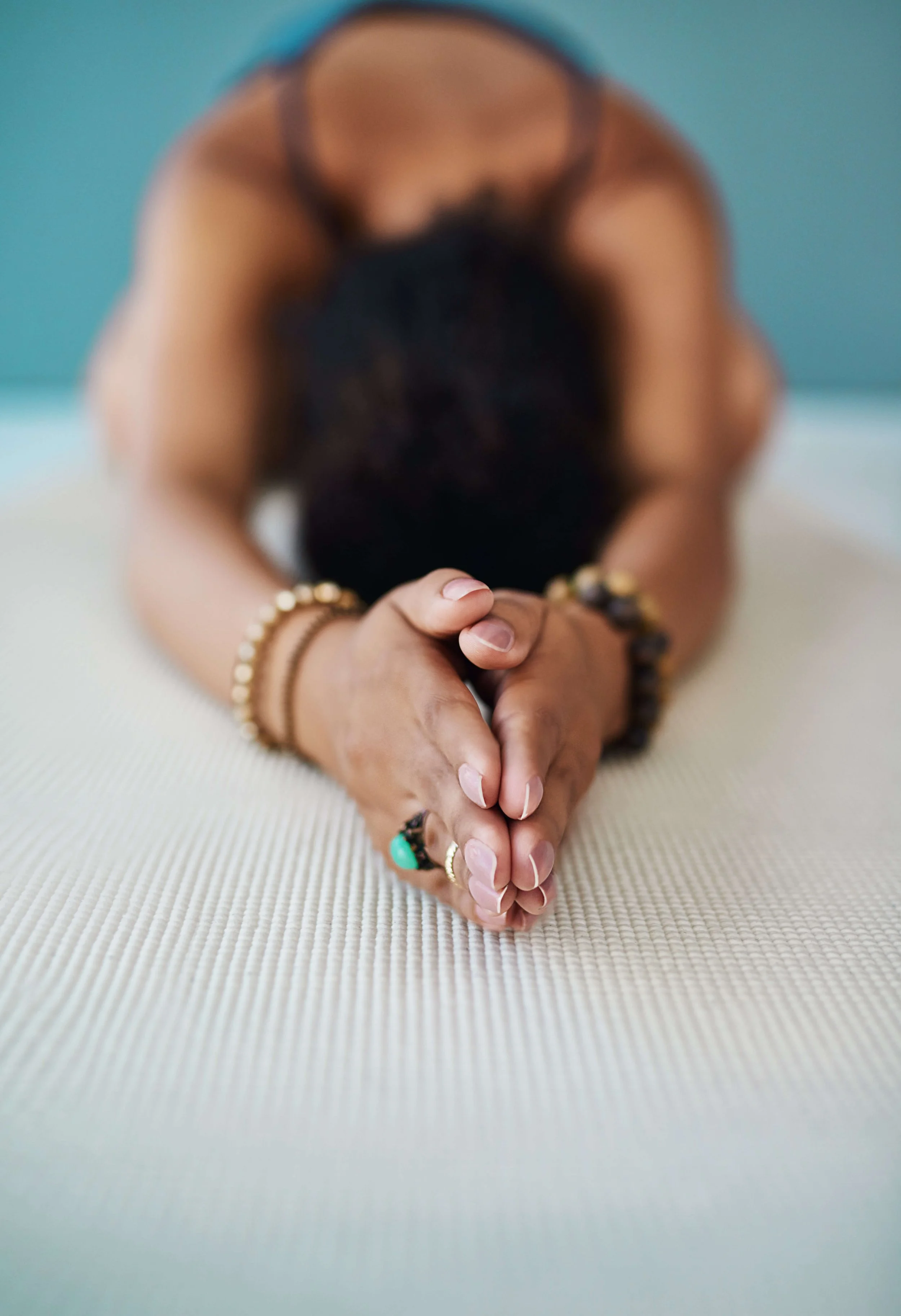A person with dark hair, wearing bracelets and rings, in a yoga pose on a light-colored mat with a teal wall background.