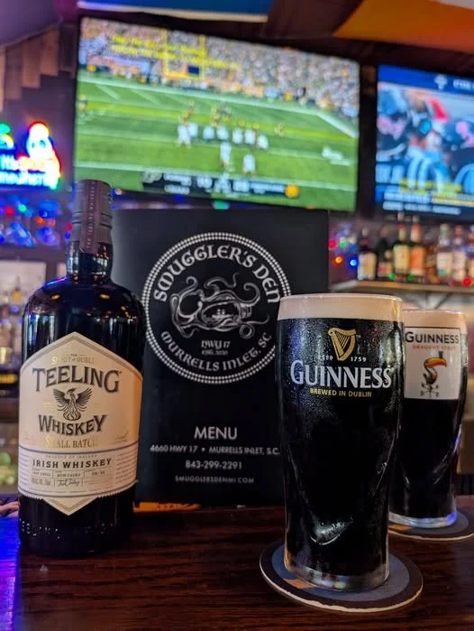 A bar counter at Smuggler's Den in Murrells Inlet SC with a bottle of Teeling Irish whiskey, a glass of Guinness beer, and another glass of Guinness behind it, near Myrtle Beach, SC