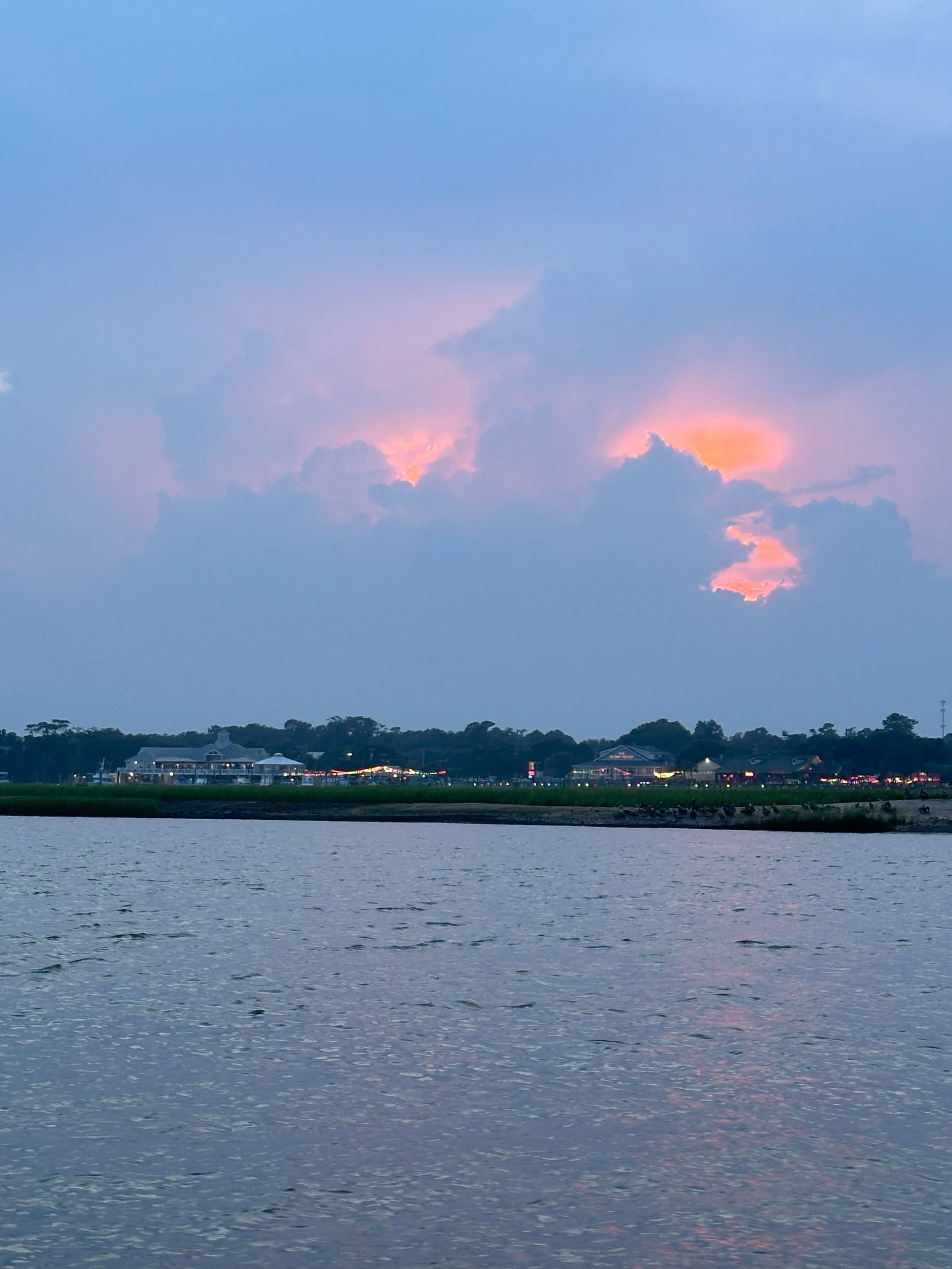 A scenic view of the marsh and the Marshwalk in Murrells Inlet, SC at sunset with a cloudy sky and some buildings and trees in the distance, near Myrtle Beach, SC
