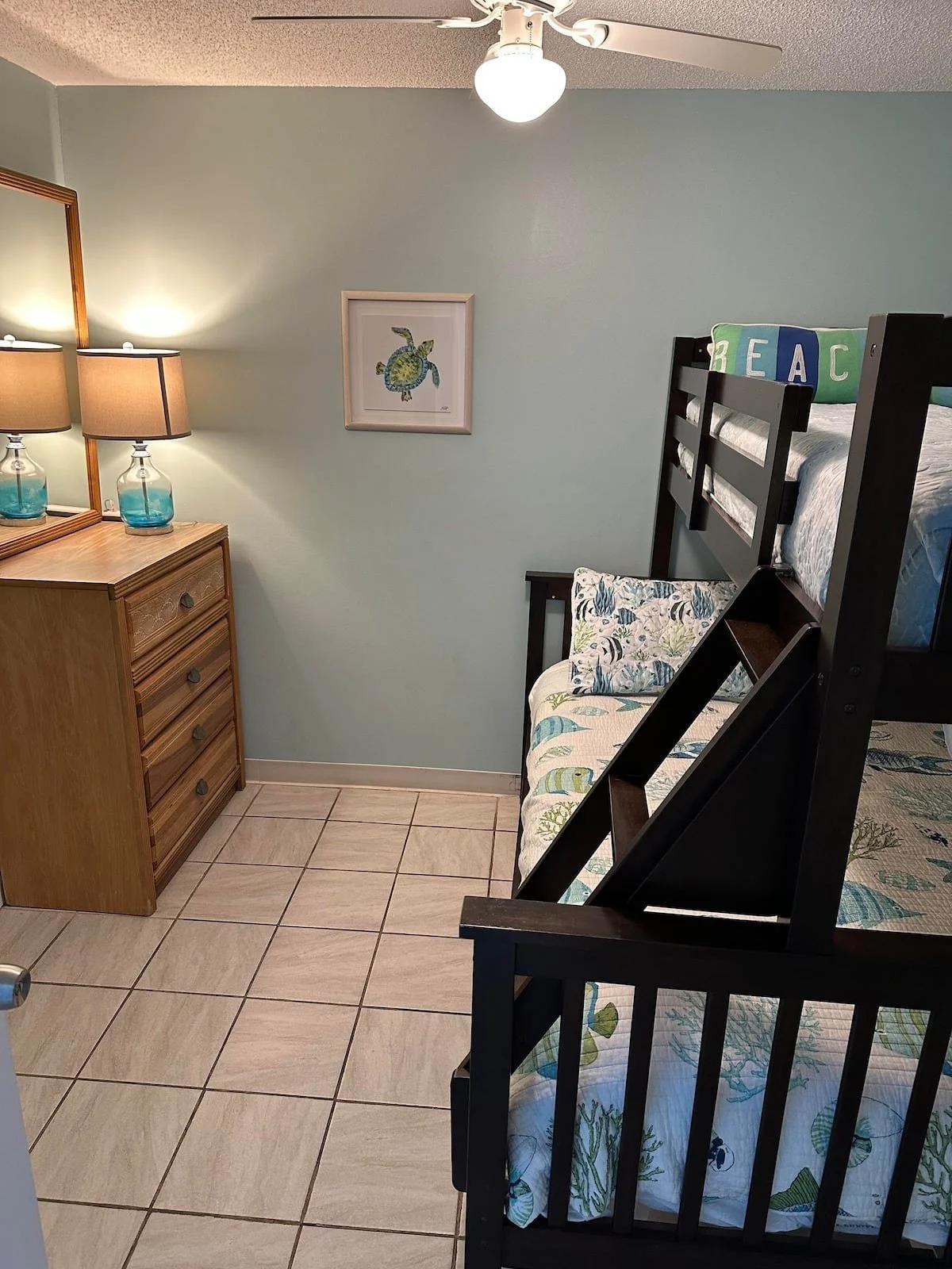 A cozy bedroom with a black bunk bed, light sea foam green wall, beige tiled floor, wooden dresser with two lamps, a framed turtle picture, and a pillow with the word 'BEACH' on it.