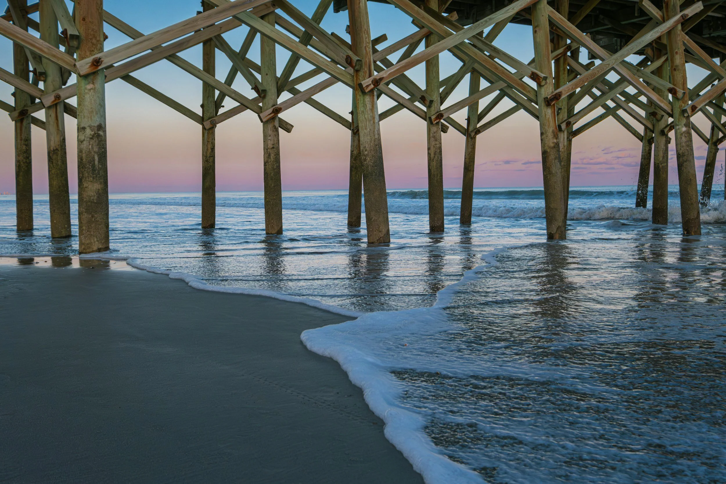 View of a beach pier with wooden support beams extending into the ocean at sunset, with gentle waves washing onto the sandy shore.