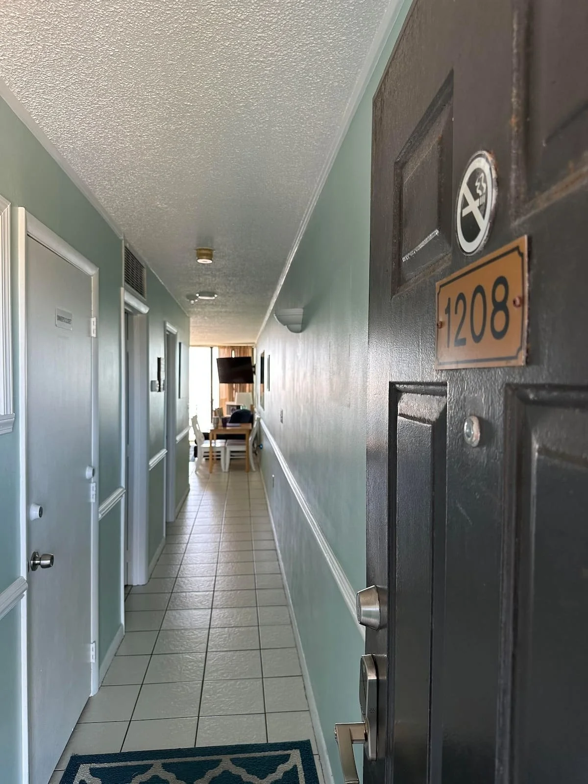 View of an apartment hallway leading to a living room with a TV and a dining table, seen from the open door of apartment 1208. The hallway has light blue walls, tiled floor, and white trim.