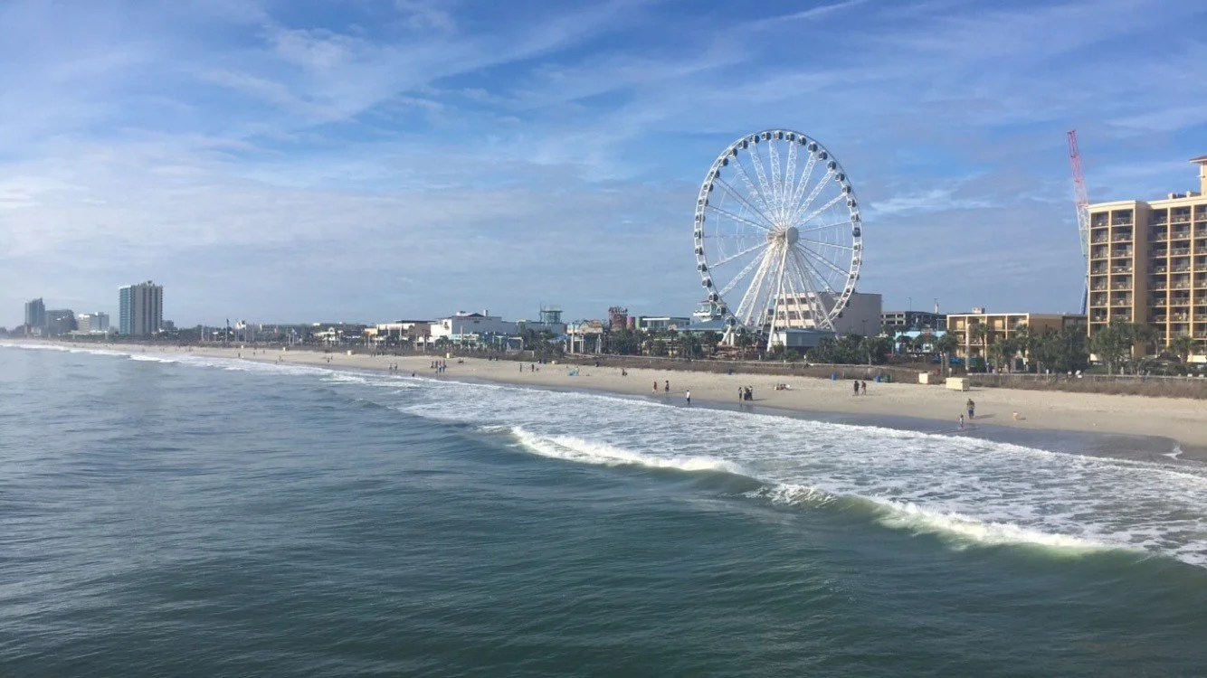 A sunny beach scene with a the Skywheel and high-rise buildings in the background, and waves rolling onto the sandy shore in Myrtle Beach, SC