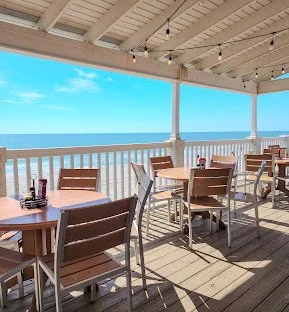 Outdoor beachside dining area at Conch Cafe with wooden tables and chairs under a covered patio, overlooking the ocean on a bright, sunny day in Garden City, SC near Myrtle Beach, SC
