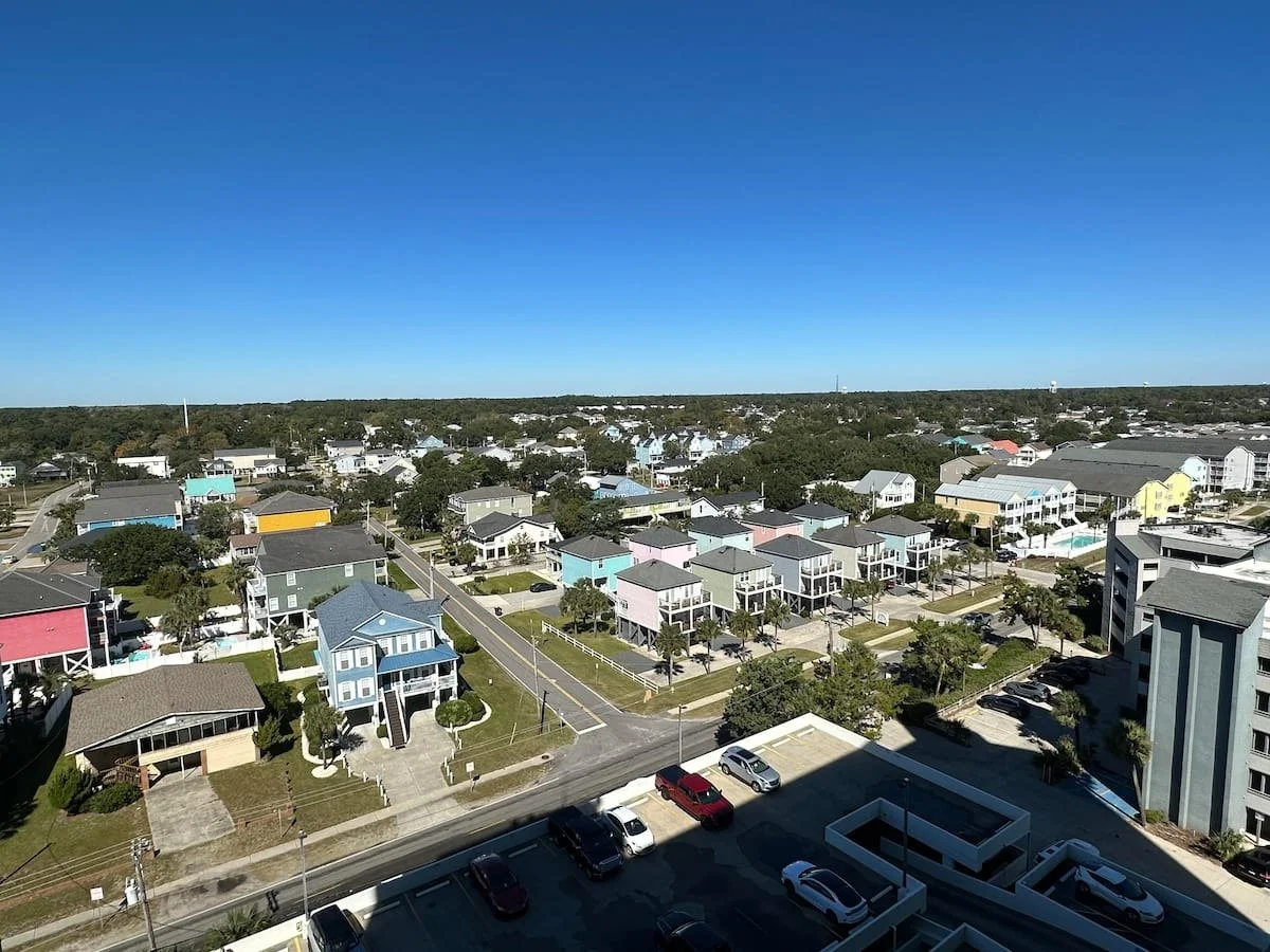 Aerial view of a seaside neighborhood with colorful beach houses, parking lot, and trees under a clear blue sky.