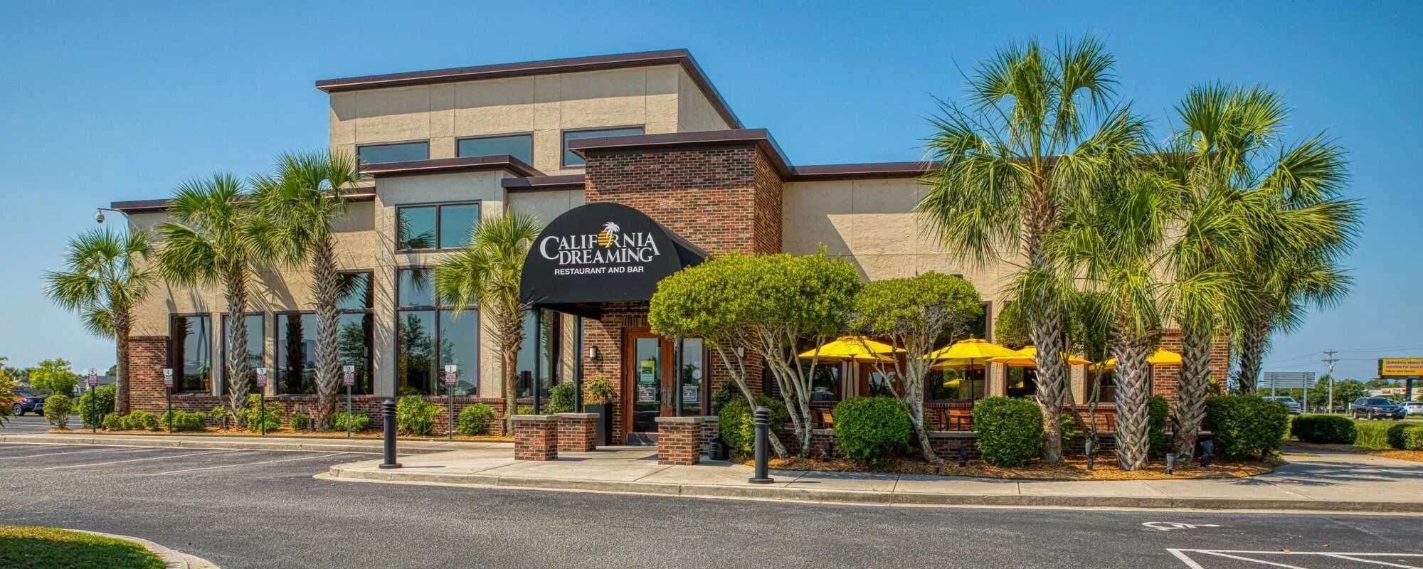 Exterior of a restaurant called California Dreaming with palm trees, bushes, outdoor seating with yellow umbrellas, and a black awning in Murrells Inlet, SC near Myrtle Beach, SC