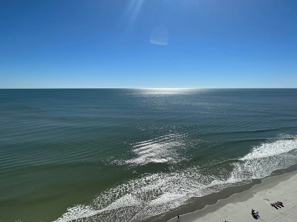 A clear view of the ocean with gentle waves, bright sunlight reflecting on the water, and a sandy beach with a few people relaxing.