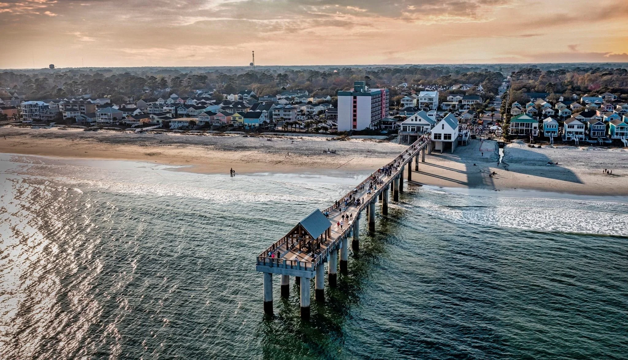 Surfside Pier extending over the ocean with people walking on it, leading to a beach and a colorful neighborhood of houses and buildings along the coast at sunset near Myrtle Beach, SC