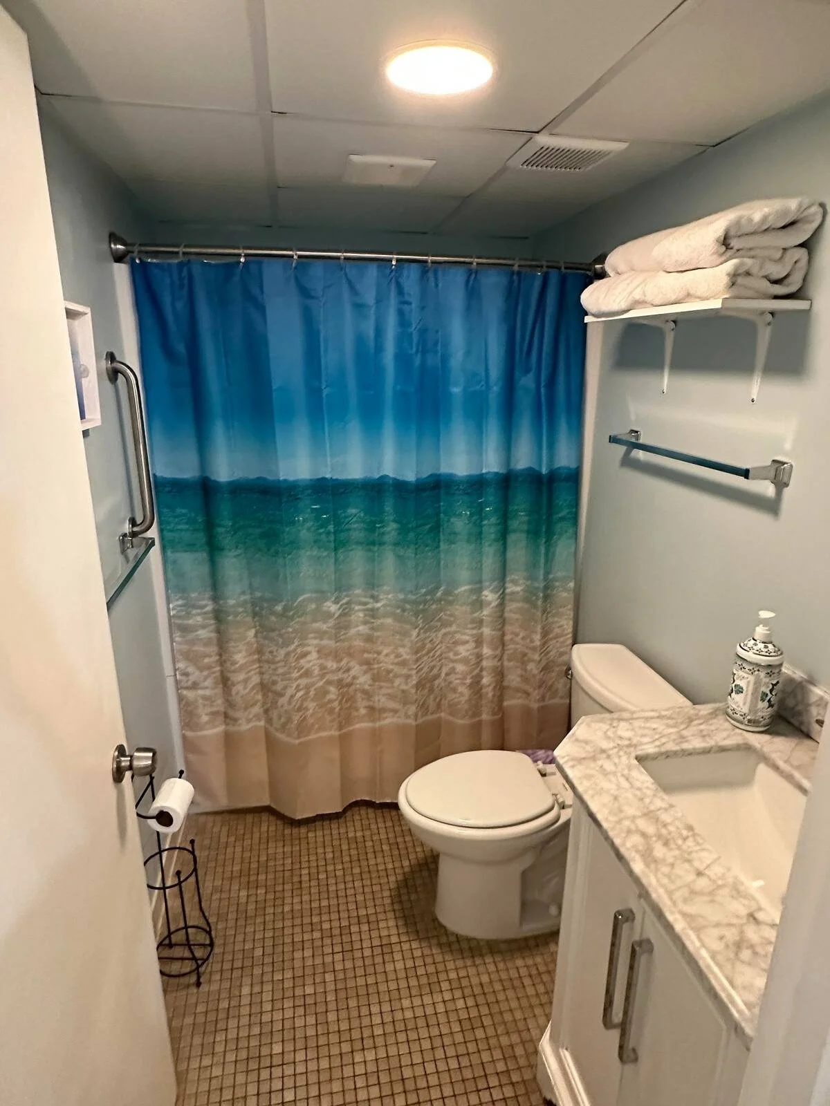 Small bathroom with a blue and beach-themed shower curtain, toilet, marble countertop sink, folded towels on a wall shelf, and beige tiled floor.
