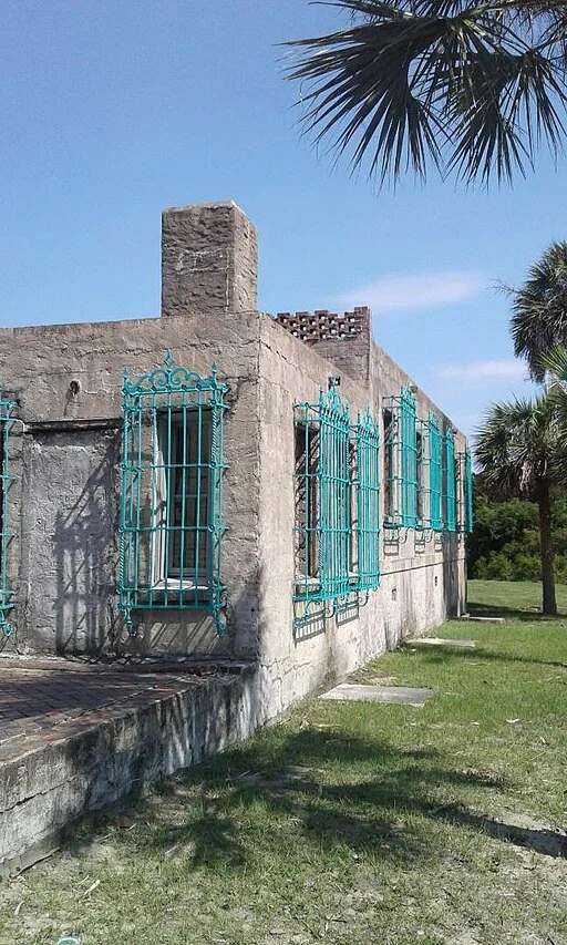 A concrete building with teal wrought iron window grilles, surrounded by grassy lawn and palm trees under a clear blue sky, Atalaya at Huntington Beach State Park in Myrtle Beach, SC