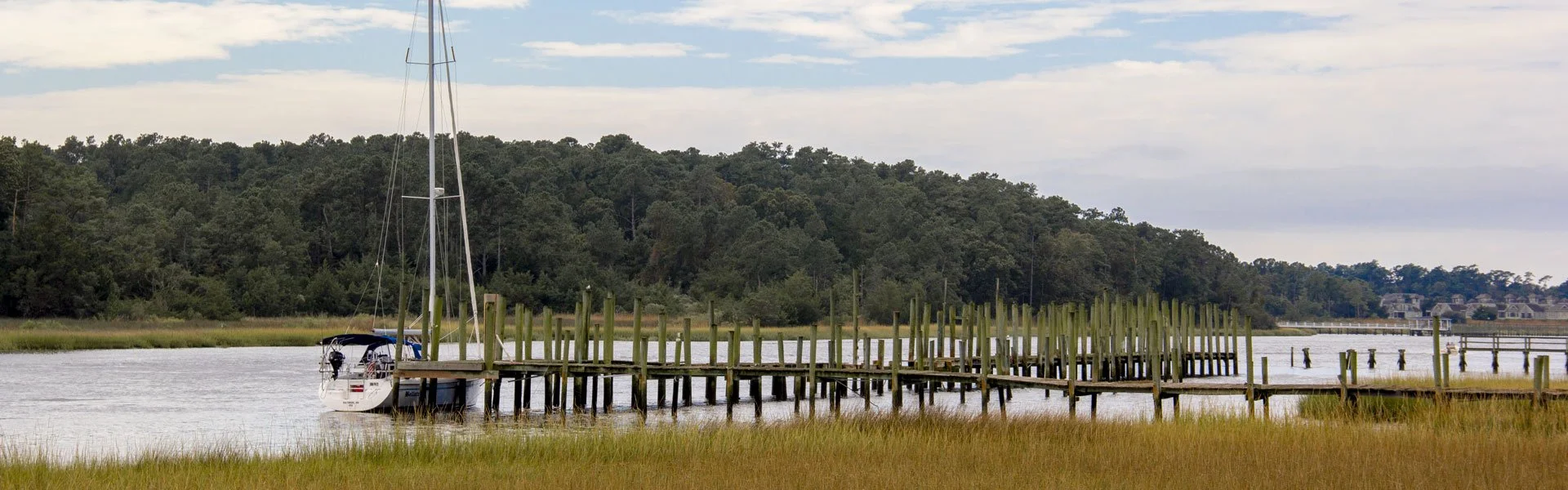 A boat dock extends into a calm water body in Little River, SC. The scene is surrounded by grassy marshland and a forested hillside in the background under a partly cloudy sky near Myrtle Beach, SC