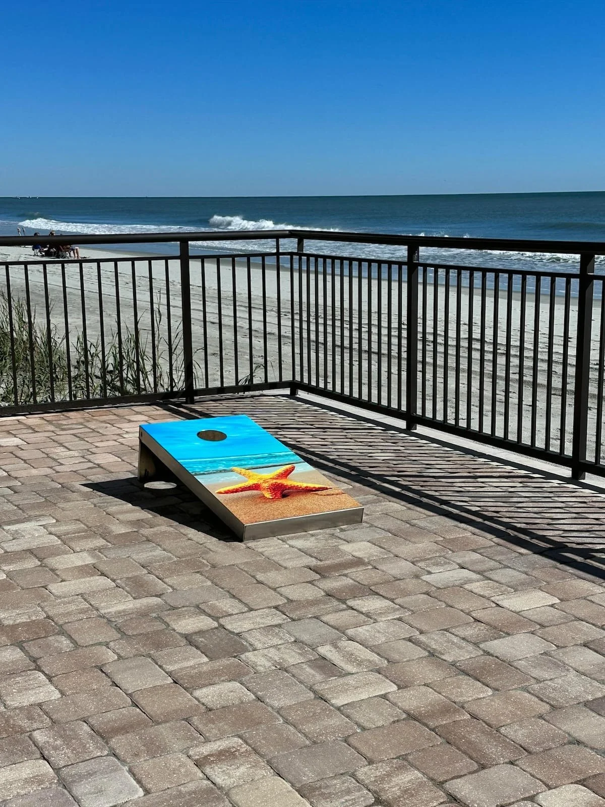A cornhole game board with a starfish painted on it, placed on a paved patio near a beach with a railing, sandy beach, ocean waves, and a clear blue sky in the background.