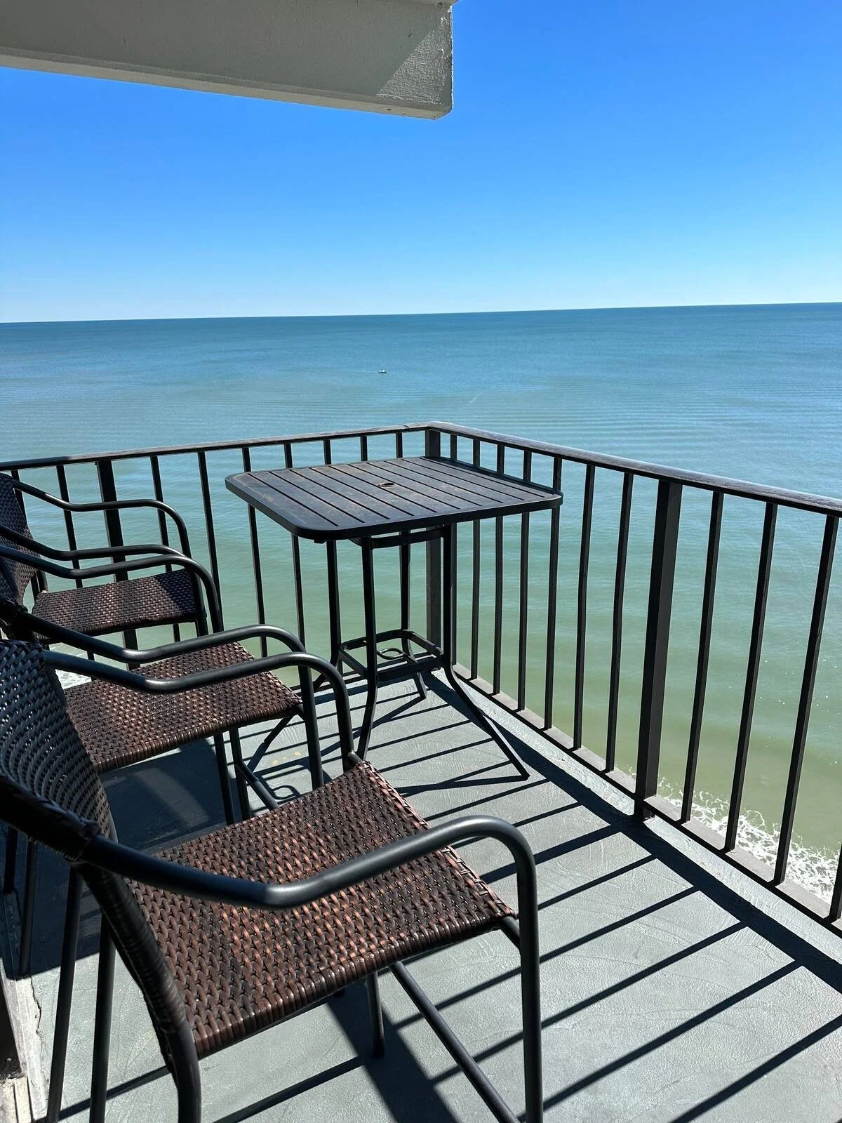 Beachside balcony with three chairs and a small table overlooking the ocean under a clear blue sky.