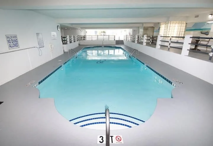 Indoor swimming pool with clear water, ladders at both ends, surrounded by white walls and a deck area, with a sign indicating pool rules.