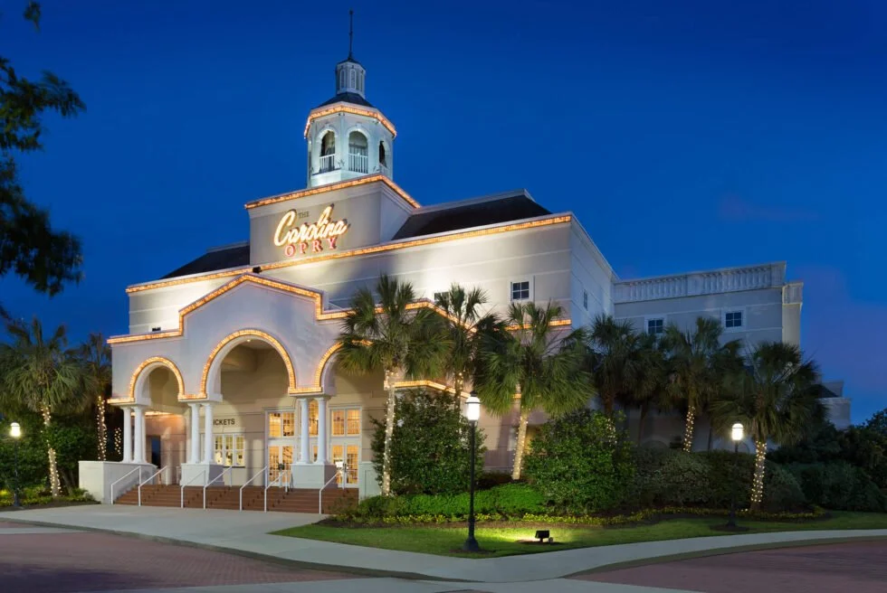 Exterior of a building with a sign that reads "The Carolina Opry," in Myrtle Beach, SC, decorated with lights, surrounded by palm trees, with a blue evening sky in the background.