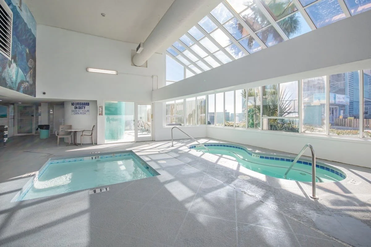 Indoor pool area with one small pool and a hot tub, large windows, sunlight, and a sign that reads 'No Lifeguard on Duty.'