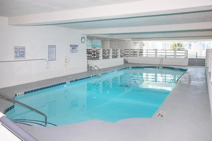 Indoor swimming pool with blue water, handrails, and lounge chairs on a deck area.