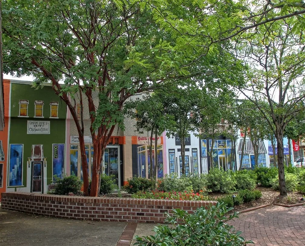 A row of small storefronts in a shopping district in Georgetown, SC, with trees and a brick planter in the foreground near Myrtle Beach, SC