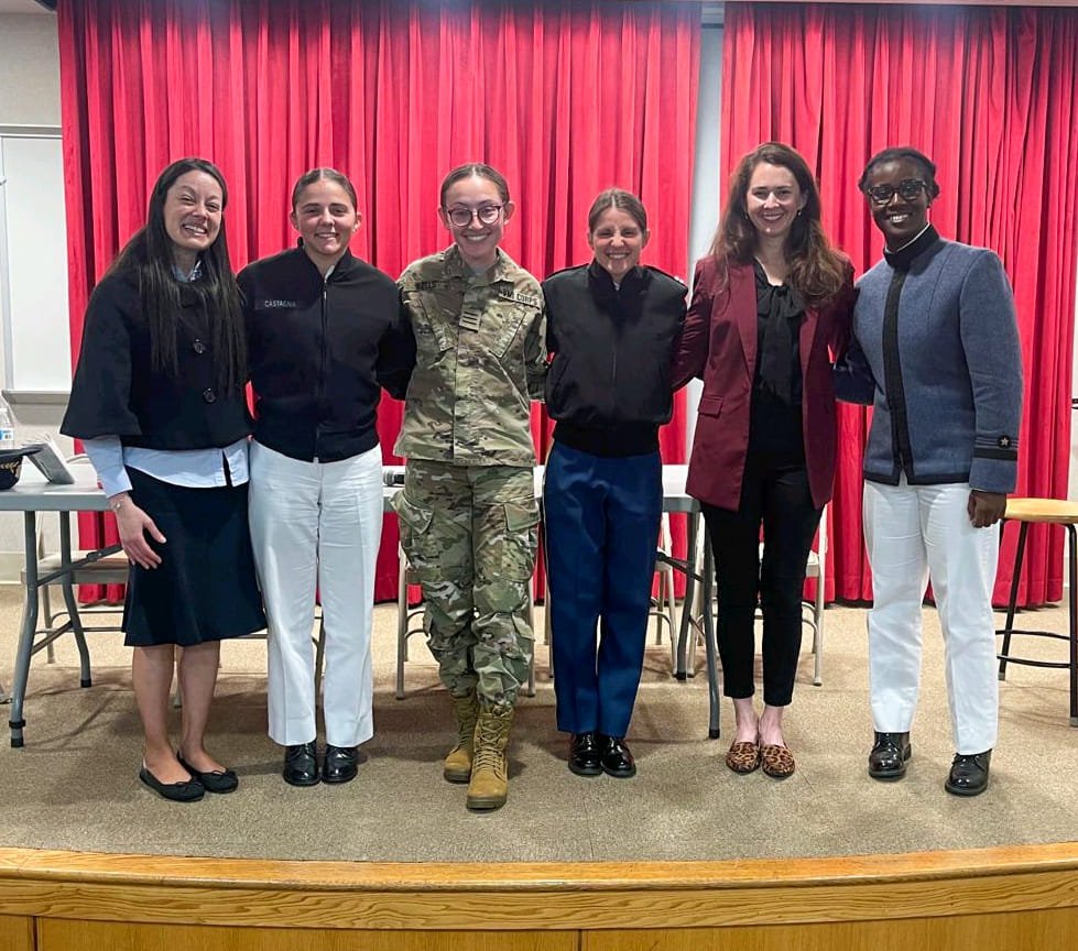 Group of six people standing in a row in front of red curtains. One person is wearing a military uniform, others in various attire.