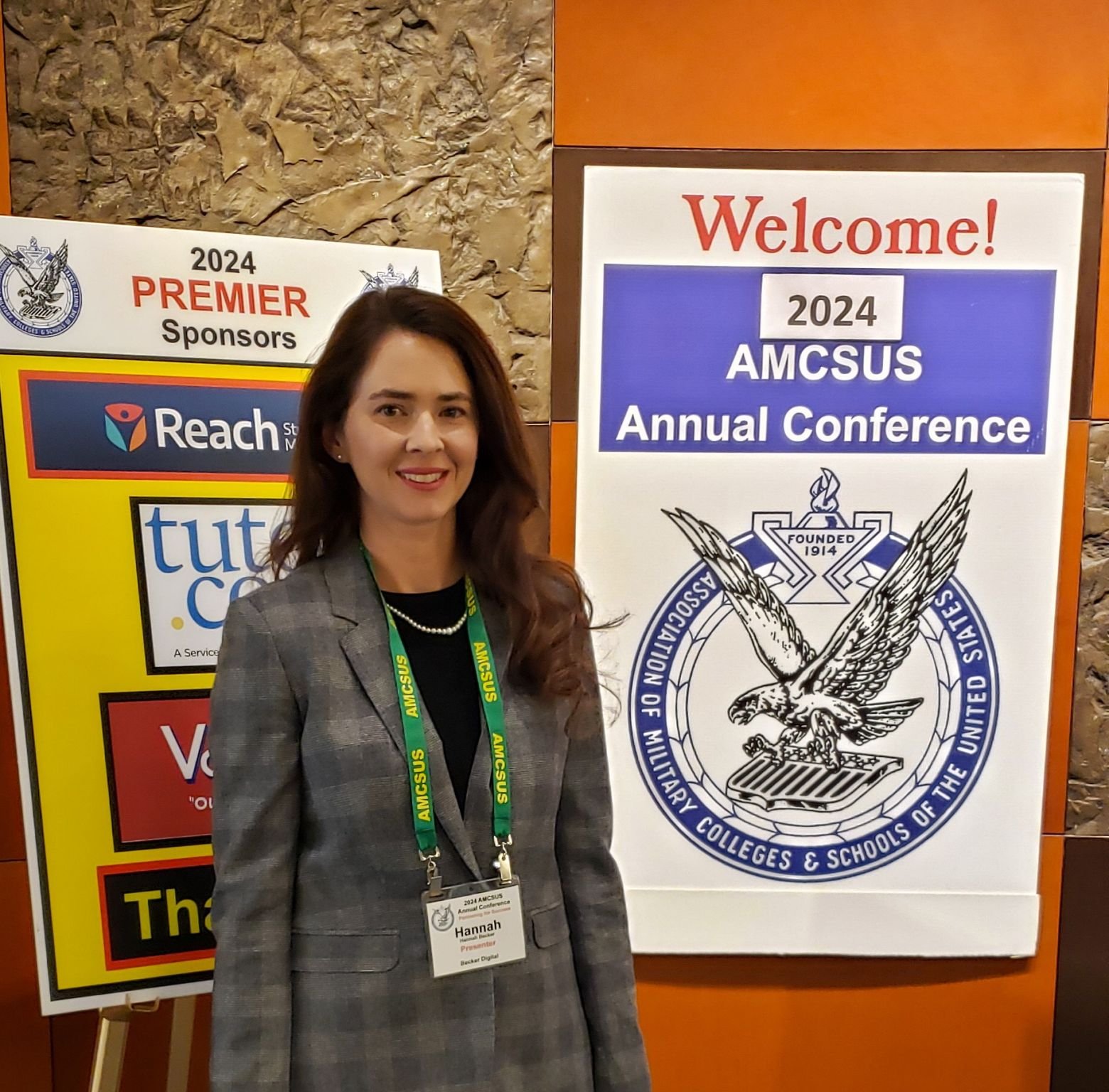 A woman standing in front of a sign for the 2024 AMCSUS Annual Conference, smiling, wearing a gray plaid blazer and a green lanyard with a name badge. Another sign behind her displays logos for event sponsors.