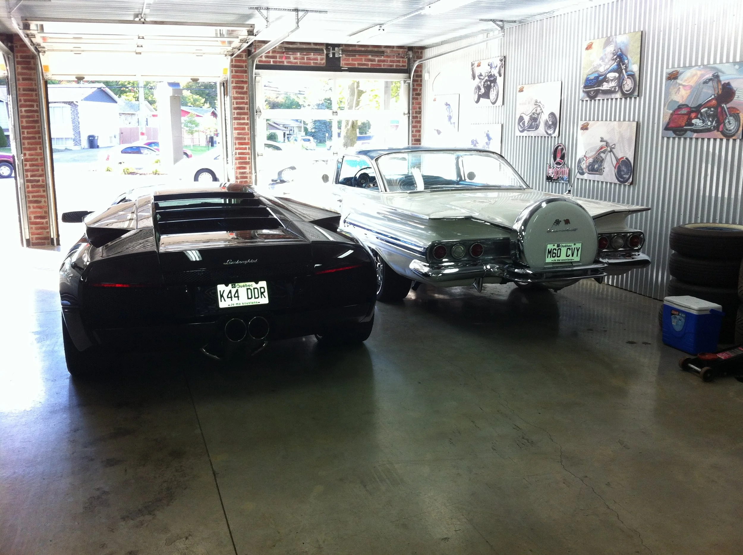 A garage with vintage and luxury cars, including a black Lamborghini and a silver Corvette, with motorcycle artwork on the walls.