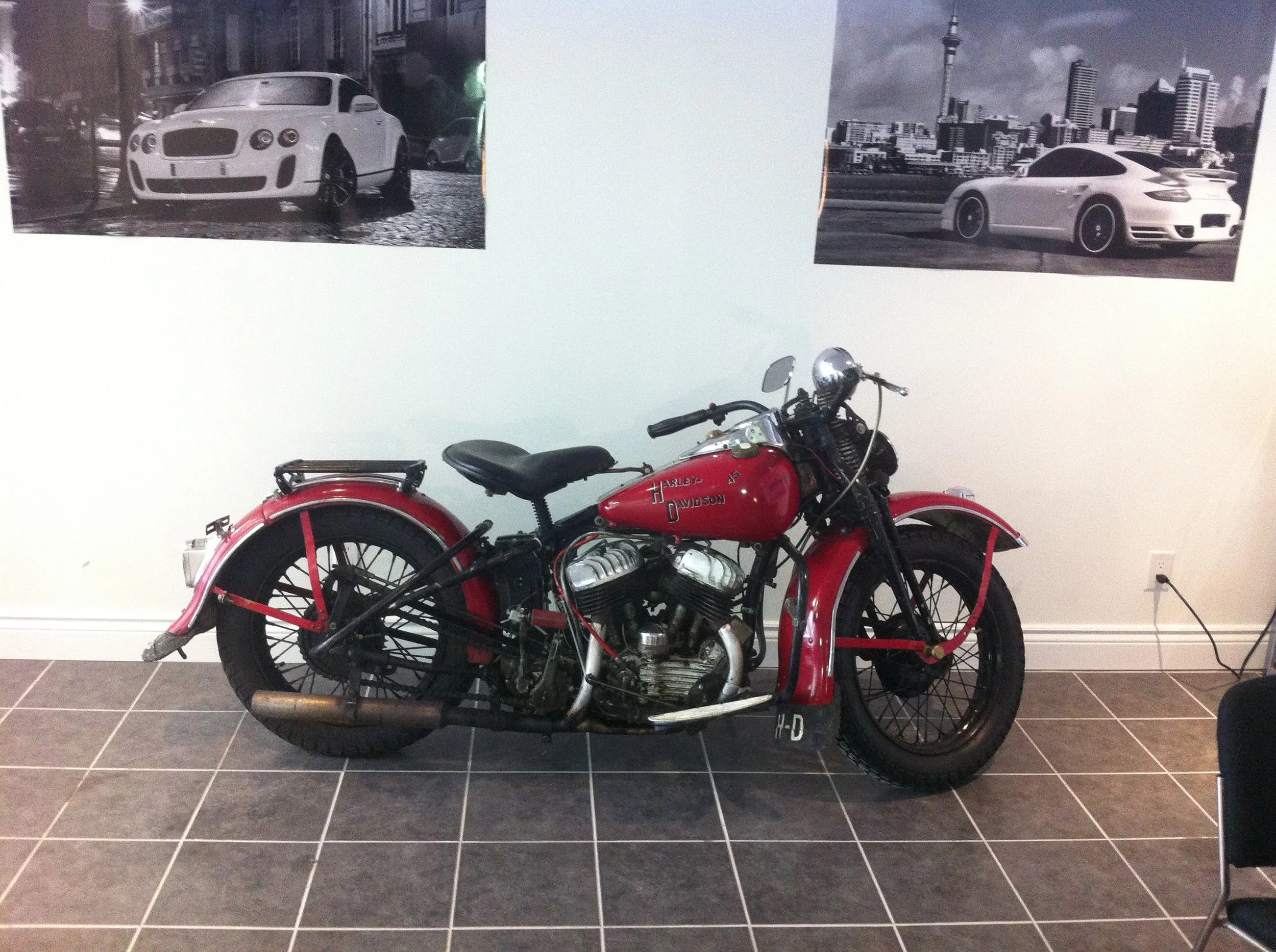 A vintage red Harley Davidson motorcycle with black accents and a black seat, parked indoors against a white wall. The wall has two black-and-white cityscape photographs hanging above the motorcycle. The floor is tiled with dark gray tiles, and there is a black rope barrier to the right.