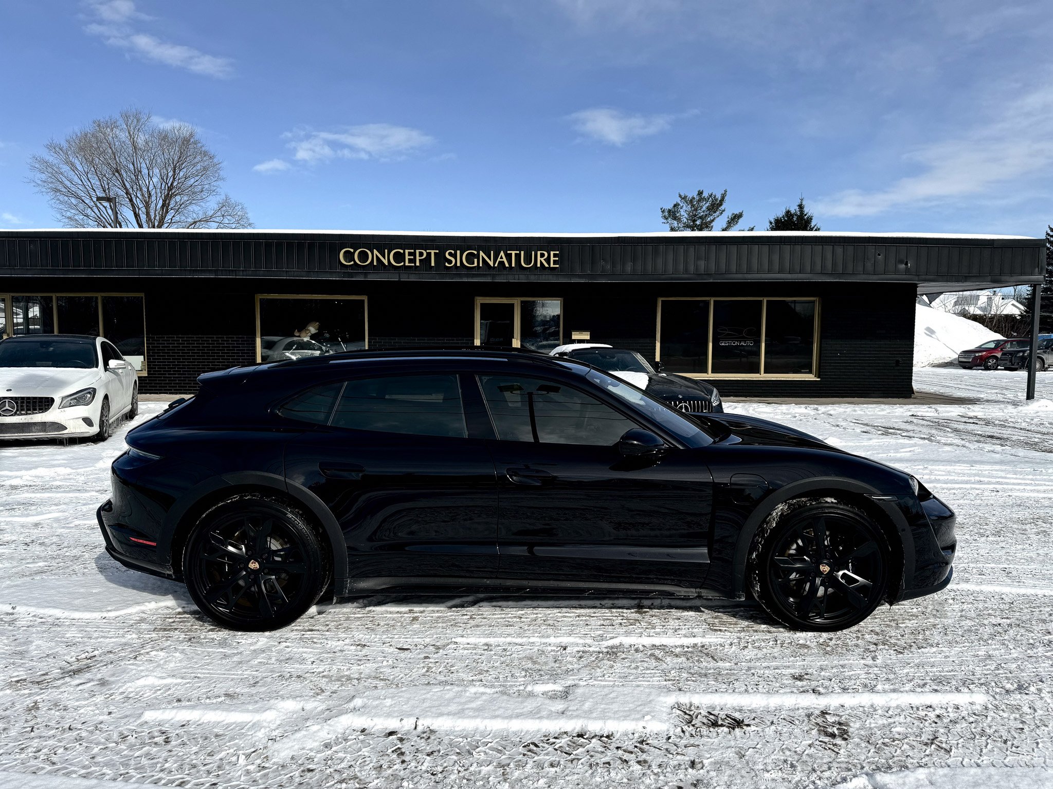 A black sports car parked in front of a black building with the sign 'CONCEPT SIGNATURE,' with other cars and snow on the ground and a partly cloudy blue sky.