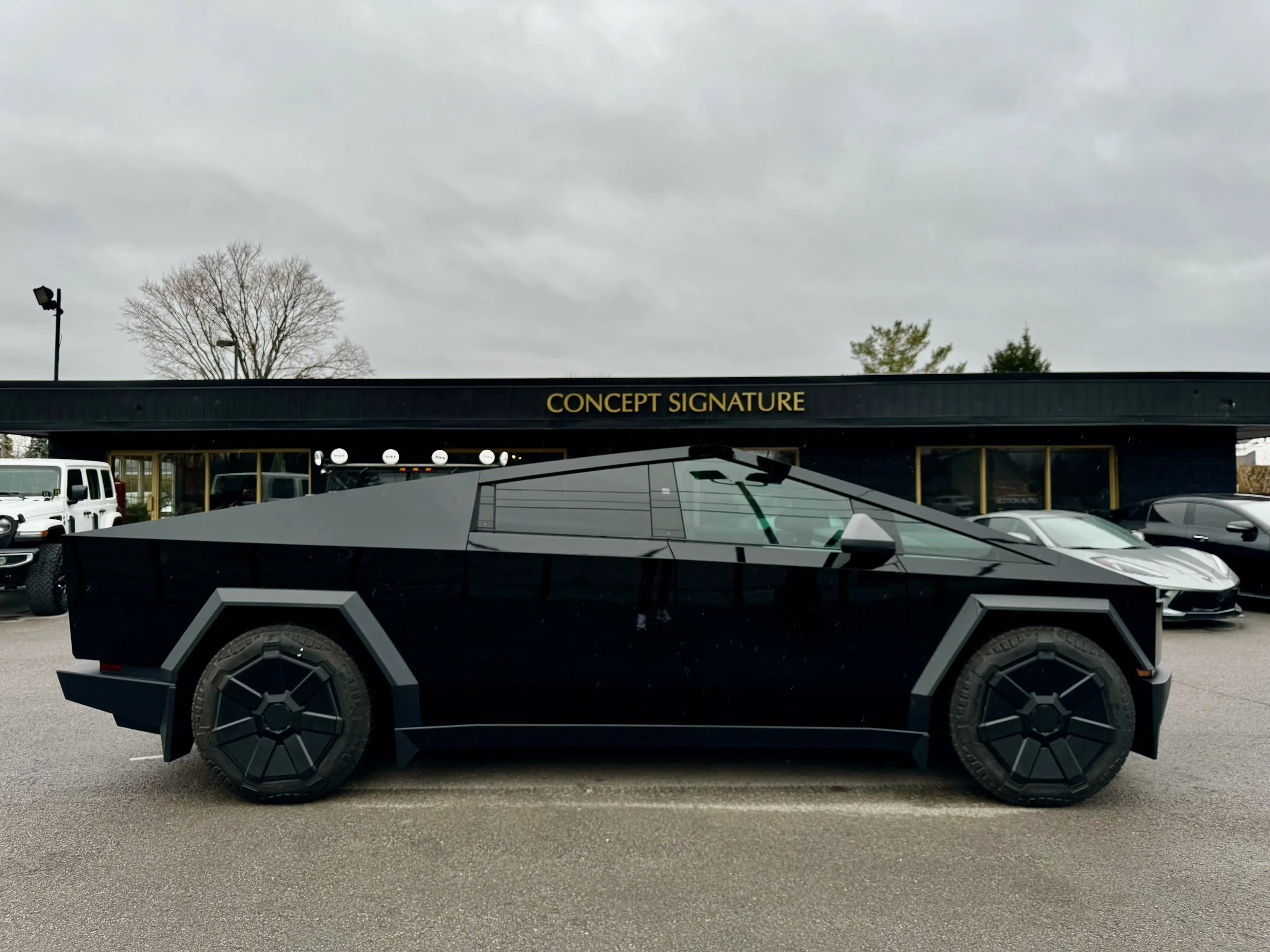 A black, futuristic-looking vehicle with angular design and large tires parked in a lot. In the background, there is a building with the sign 'Concept Signature,' a few trees, and vehicles under a cloudy sky.
