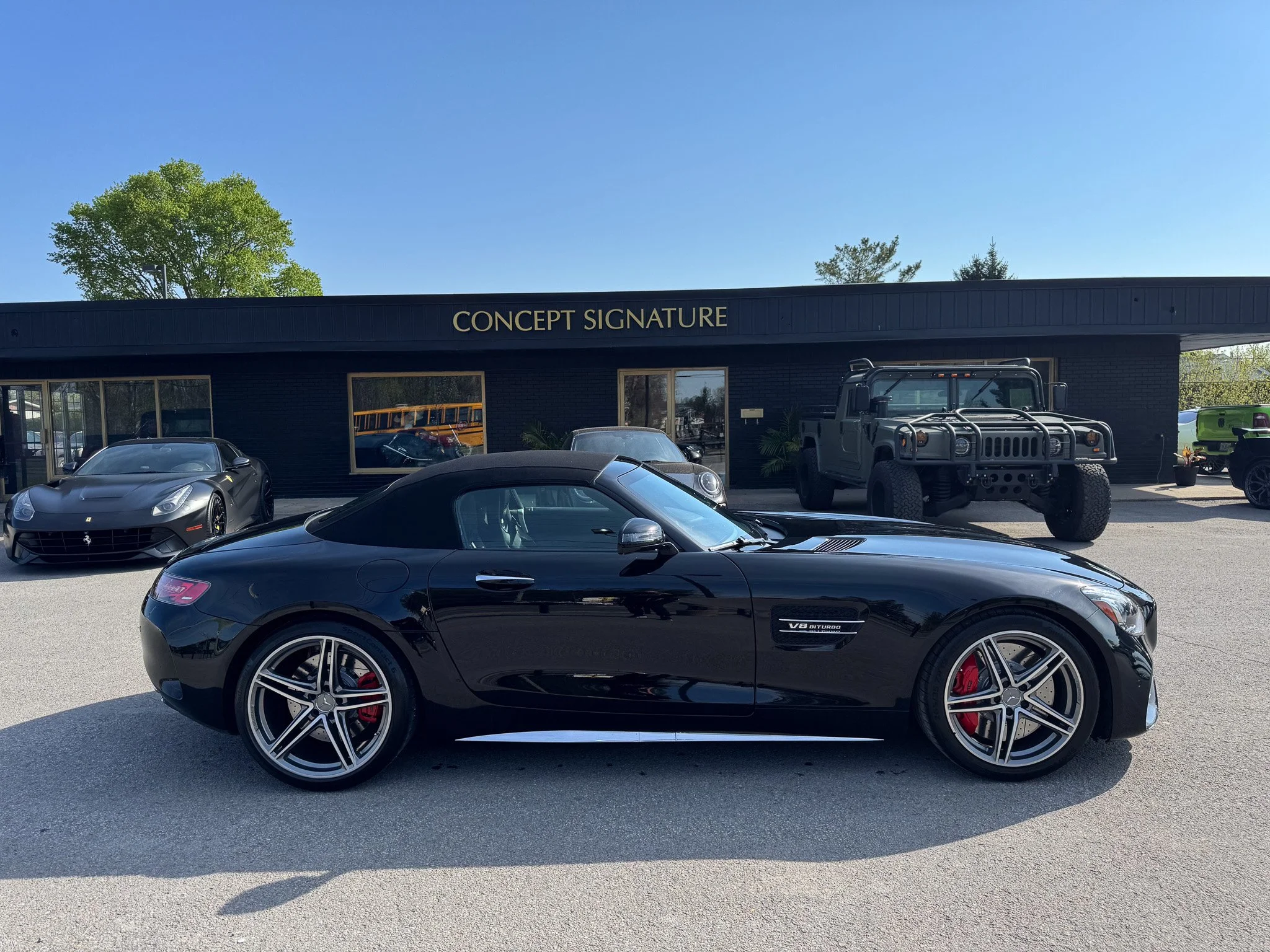 Front view of a black Mercedes-Benz sports car parked in a lot, with a commercial building labeled 'Concept Signature' and other vehicles, including a military-style truck, in the background under a clear blue sky.