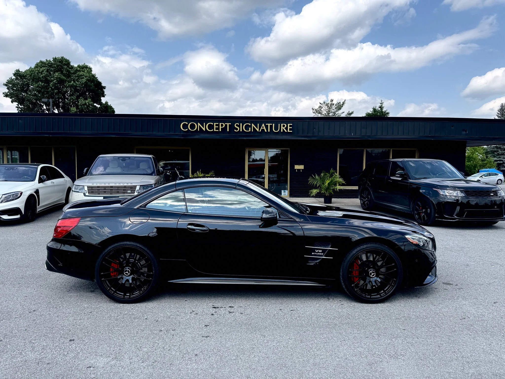 A parking lot filled with luxury cars in front of a dark building with a sign that reads 'Concept Signature.' The sky above is partly cloudy with some large trees visible in the background.