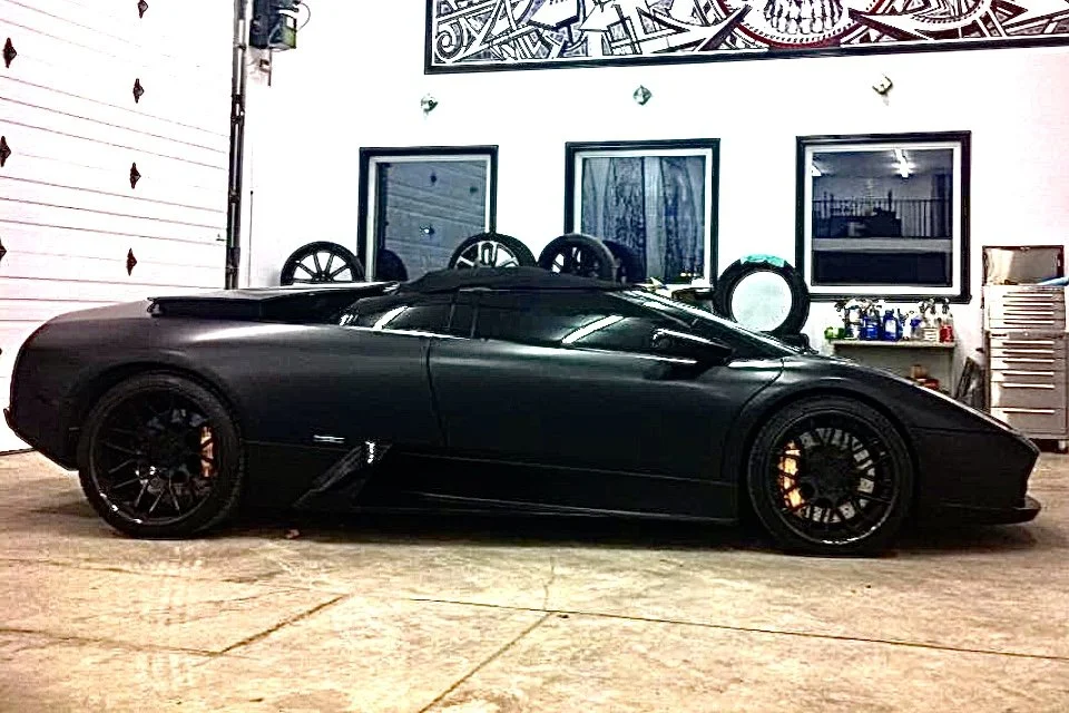 A black sports car, possibly a Lamborghini, parked inside a garage with white walls and three framed windows or artworks behind it.