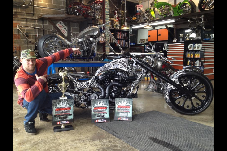 A man kneeling beside a custom motorcycle with trophies and award plaques in front of it, inside a workshop with motorcycles and tools.