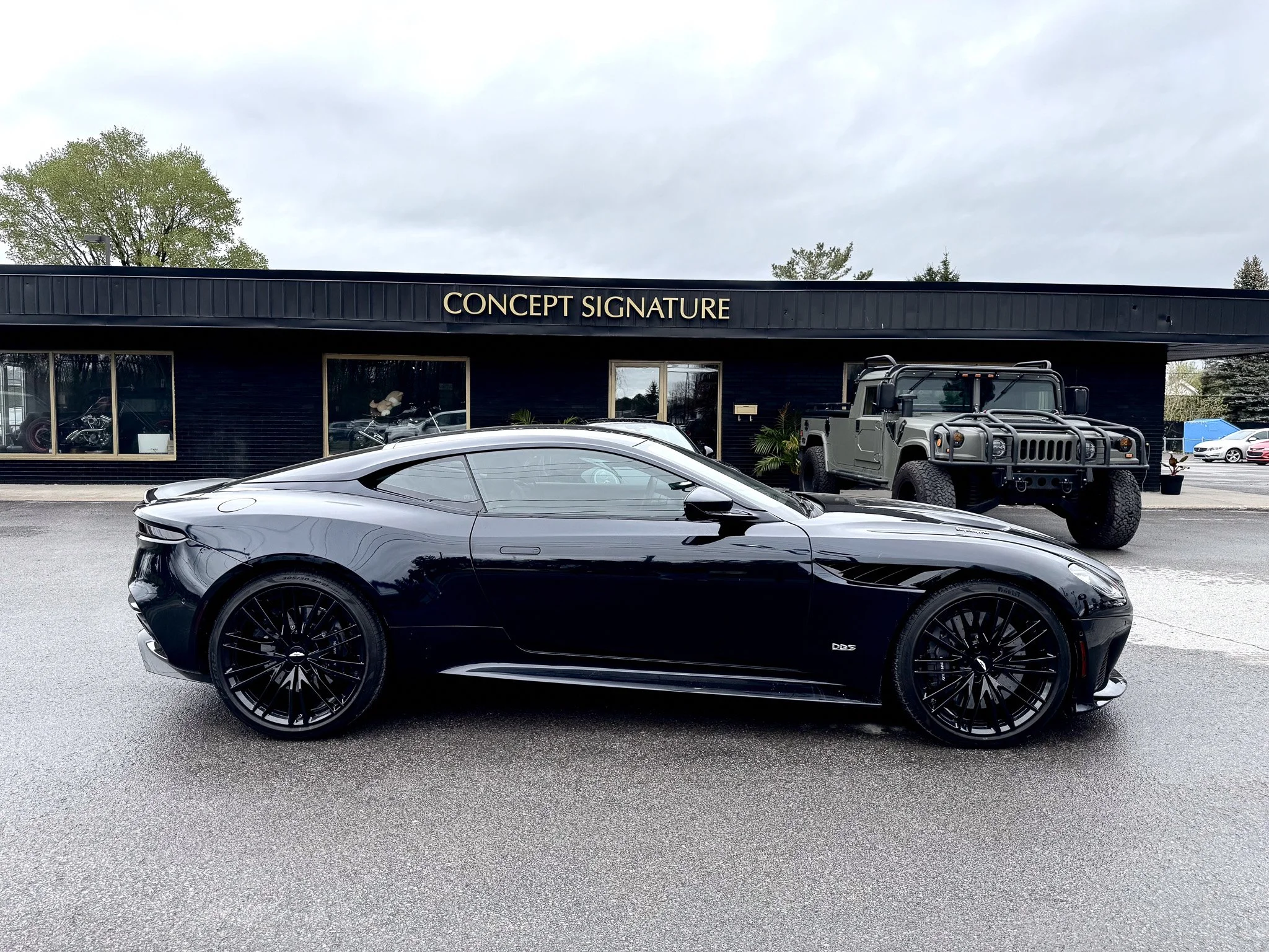A sleek black sports car parked in front of a black building with the sign 'CONCEPT SIGNATURE', alongside other vehicles including a military-style vehicle and a motorcycle inside the window.
