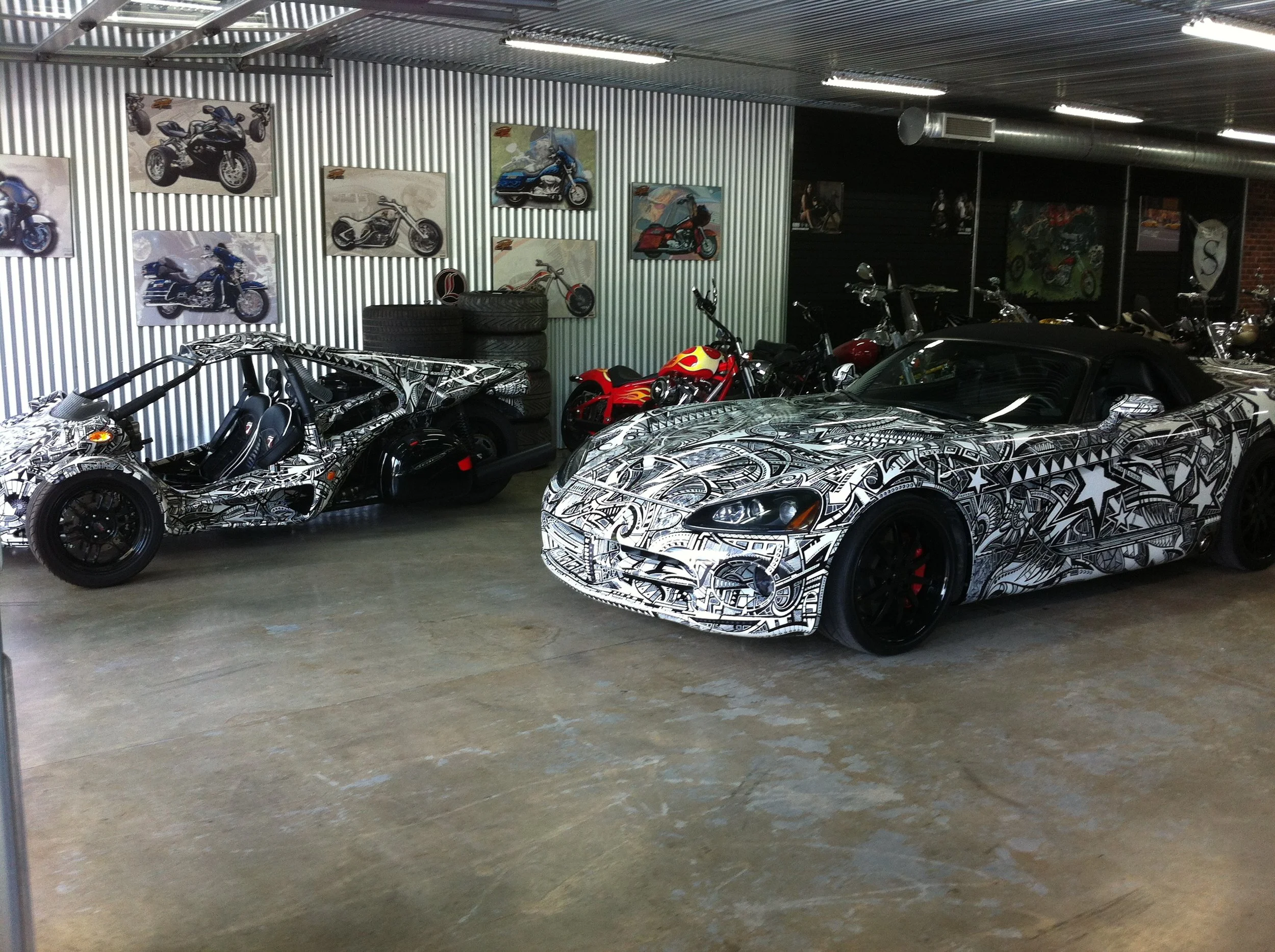 Interior of a garage with multiple motorcycles and sports cars, some displaying camouflage and graffiti-style designs; walls decorated with motorcycle posters.