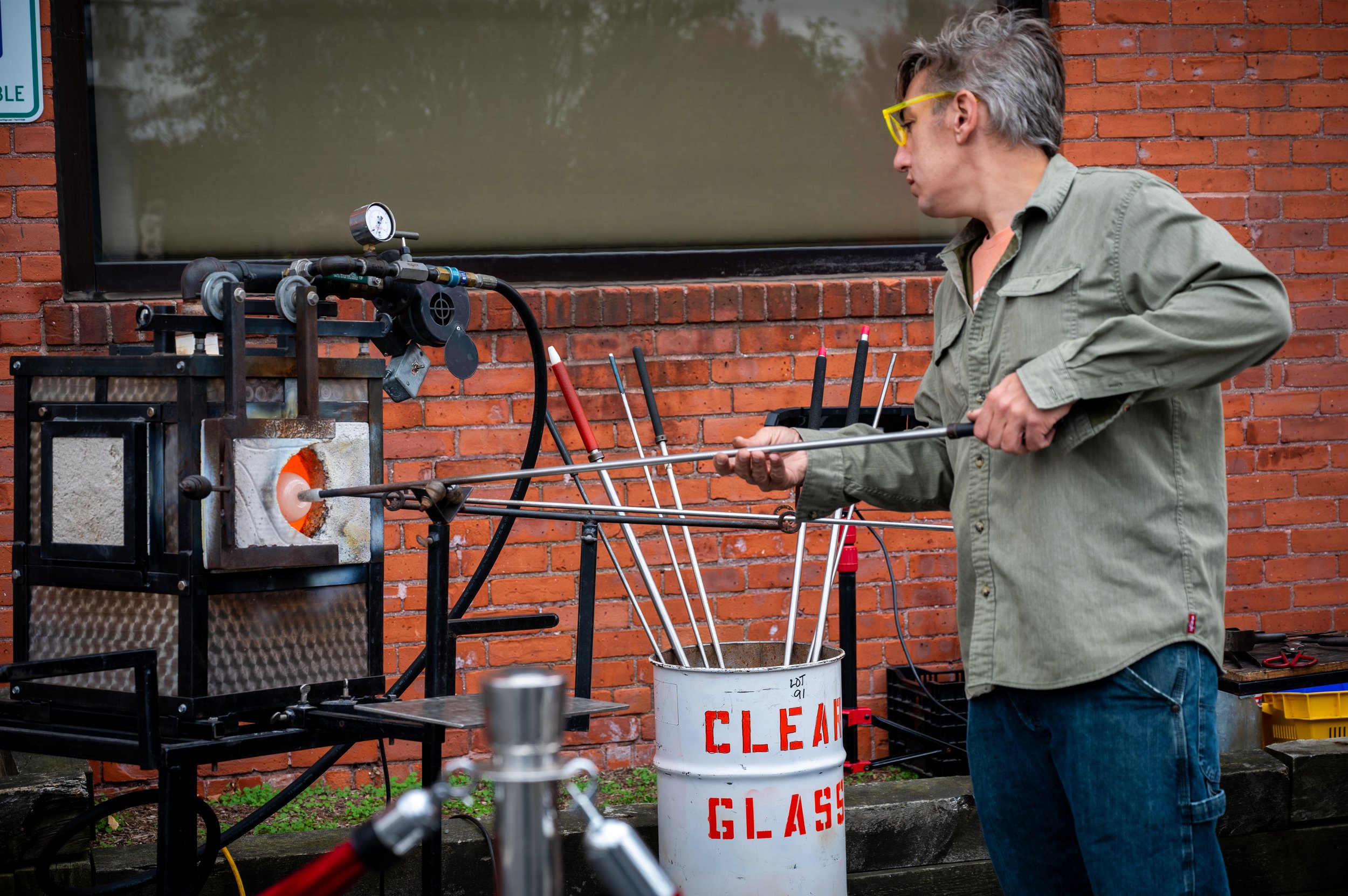 A man is glassblowing a glowing orange glass piece in a workshop with a brick wall background.