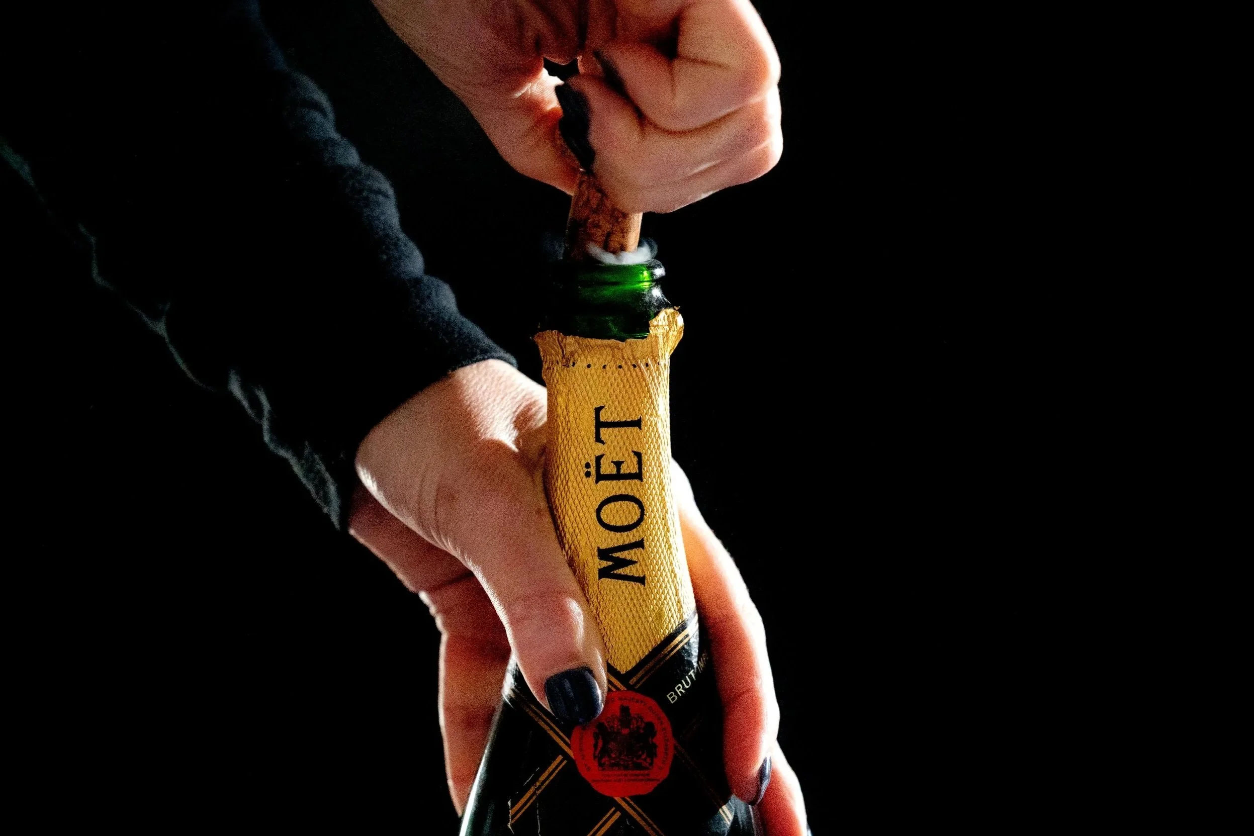A person opening a champagne bottle with a gold label, against a black background.