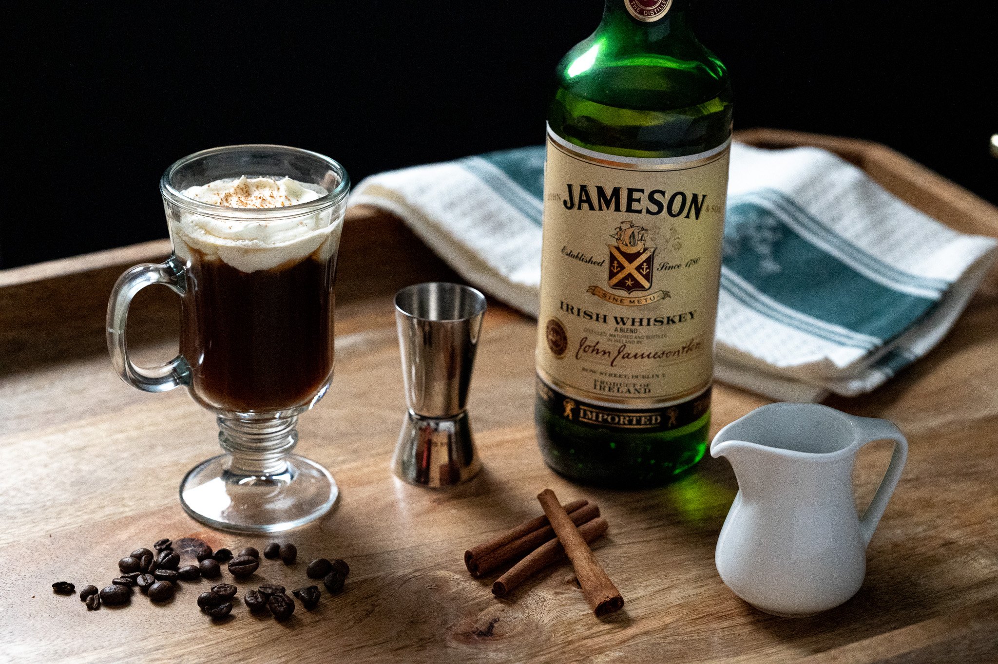 A glass of Irish coffee topped with whipped cream, a bottle of Jameson Irish whiskey, a stainless steel jigger, coffee beans, cinnamon sticks, a white creamer pitcher, and a striped kitchen towel on a wooden tray.