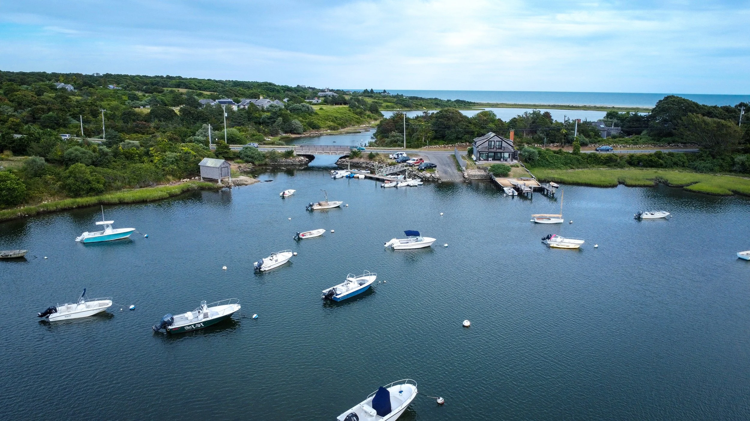 Aerial view of a small harbor with several boats anchored in calm waters, surrounded by green landscape and houses, with a road and the ocean in the background.