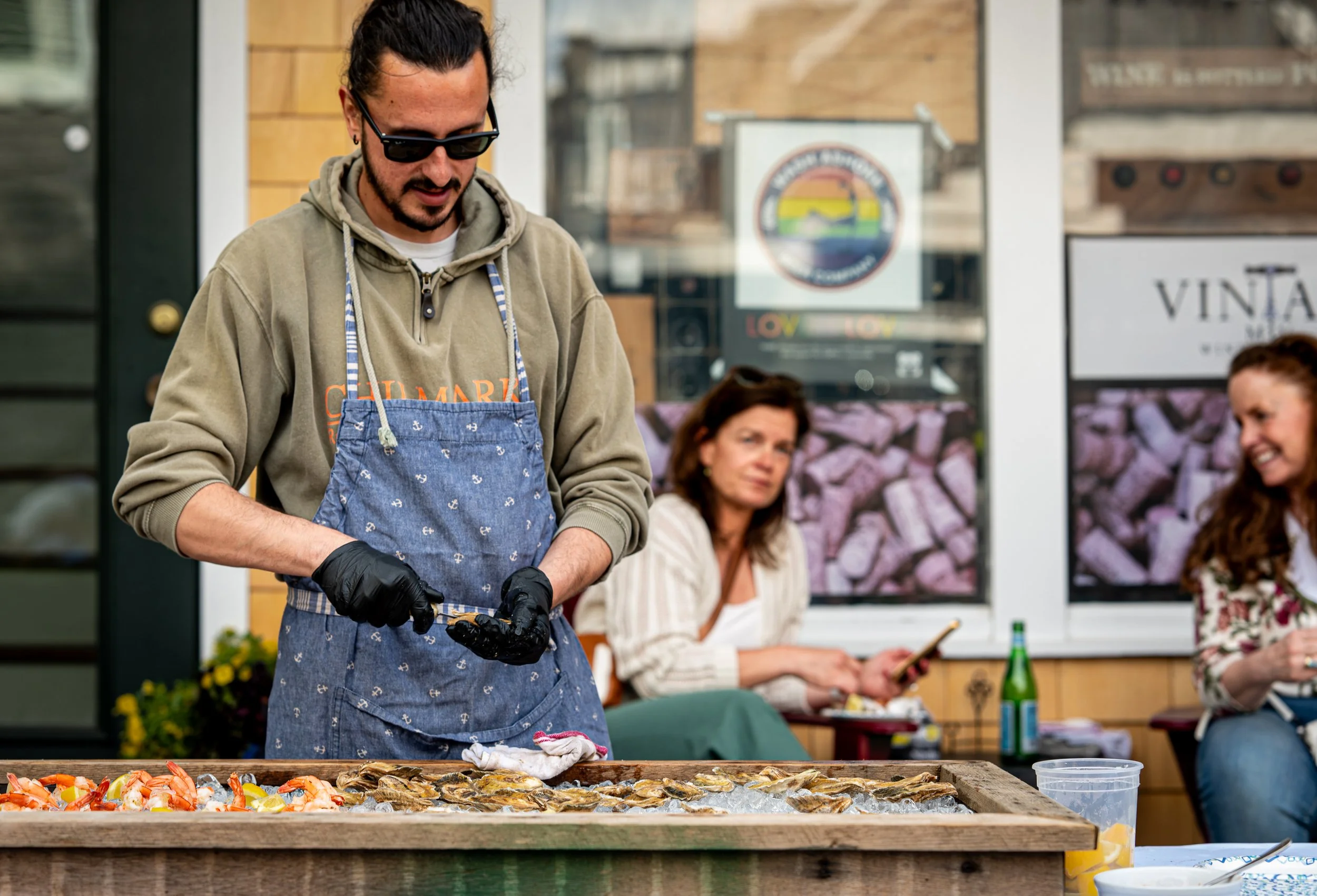Man wearing sunglasses, apron, and gloves preparing seafood at an outdoor market, with two women seated in the background, one with a smartphone and the other smiling.