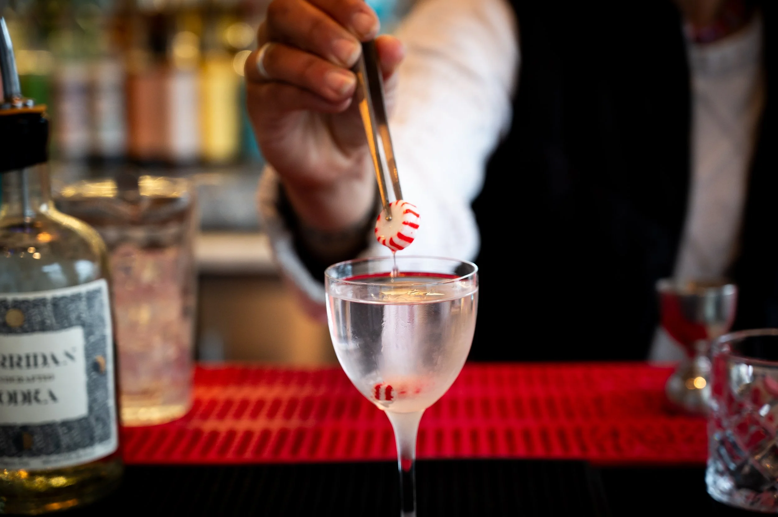 A bartender is dropping a peppermint candy into a tall glass of clear water, with bar bottles in the background.