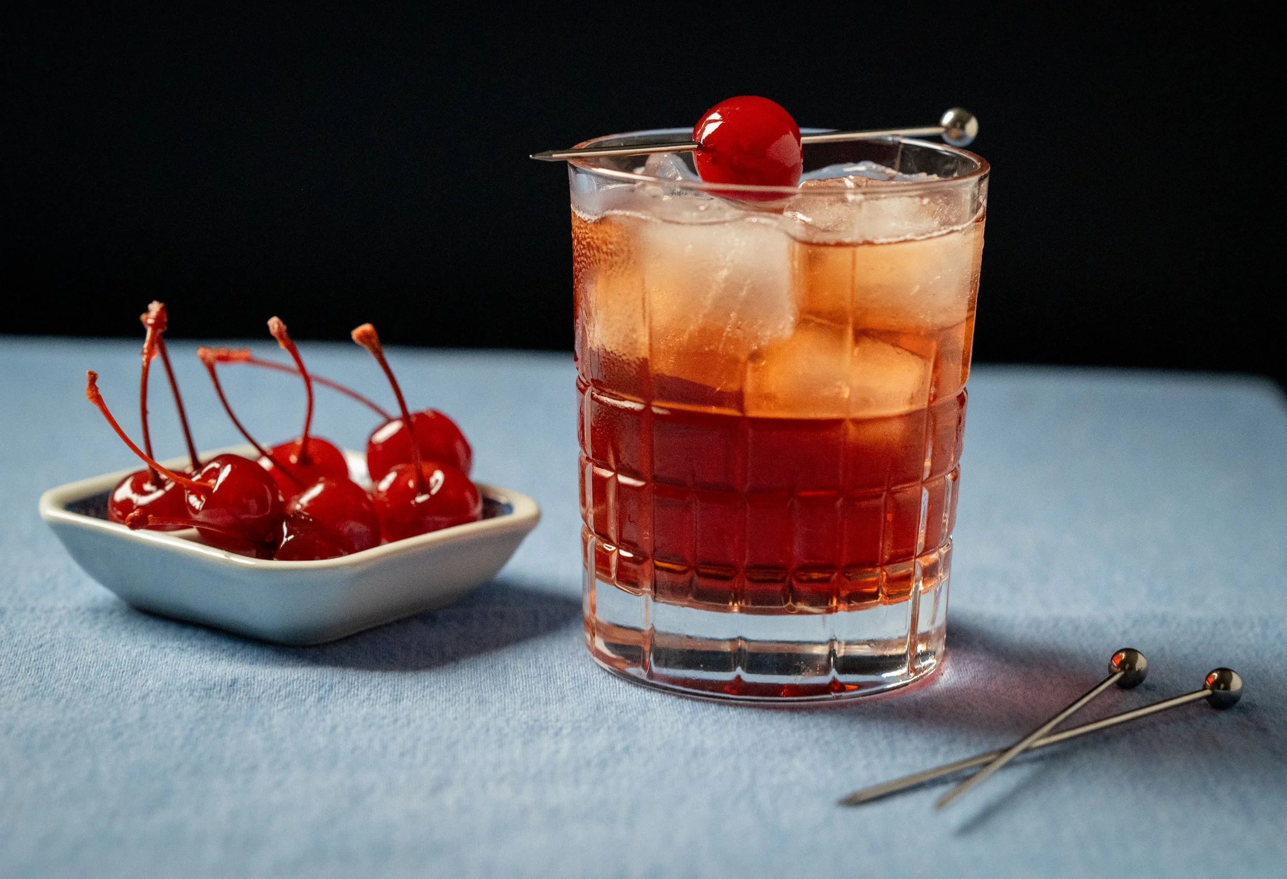 A glass of cherry cocktail with ice and a cherry garnish, next to a small dish of fresh cherries on a light blue surface with a dark background.
