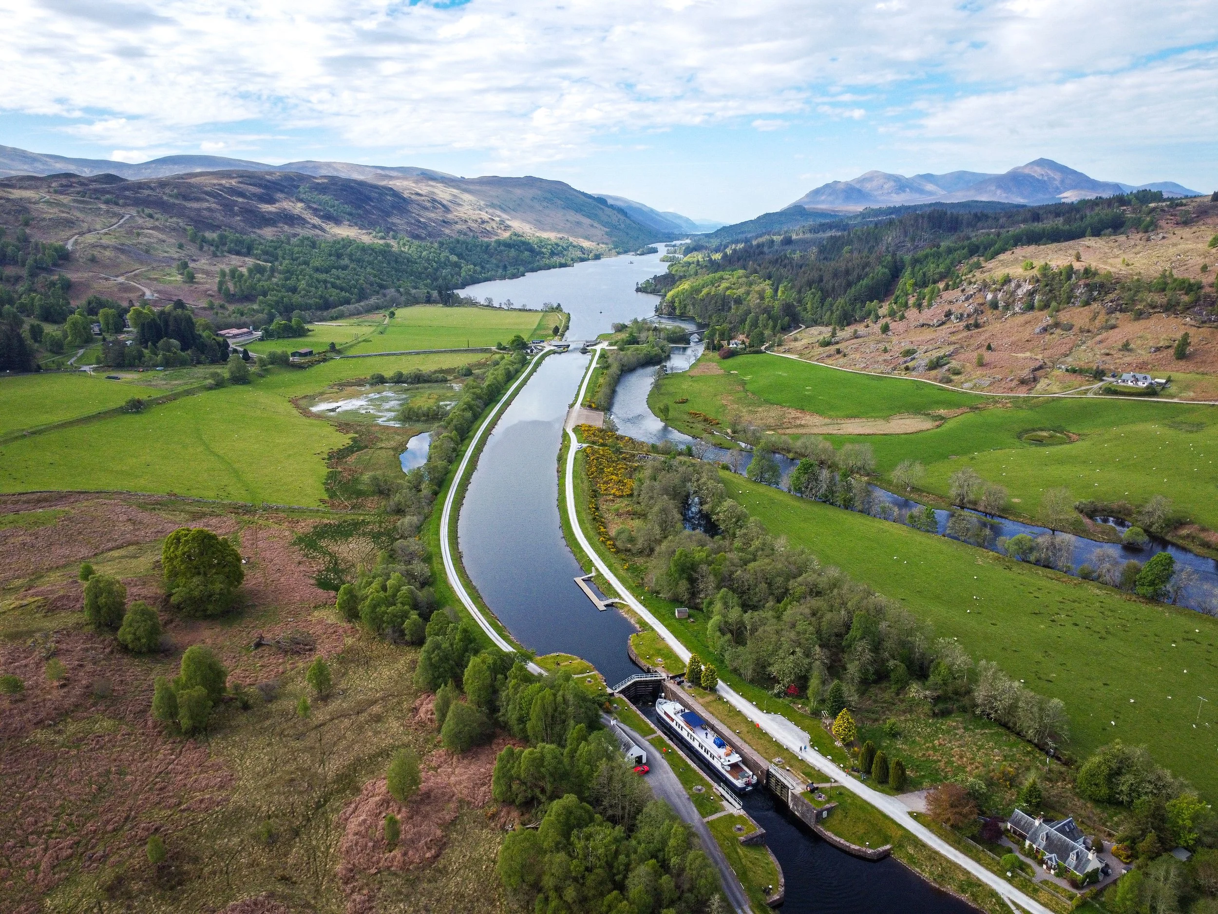 Aerial view of a winding canal and river through lush green farmland and rolling hills with mountains in the background on a partly cloudy day.