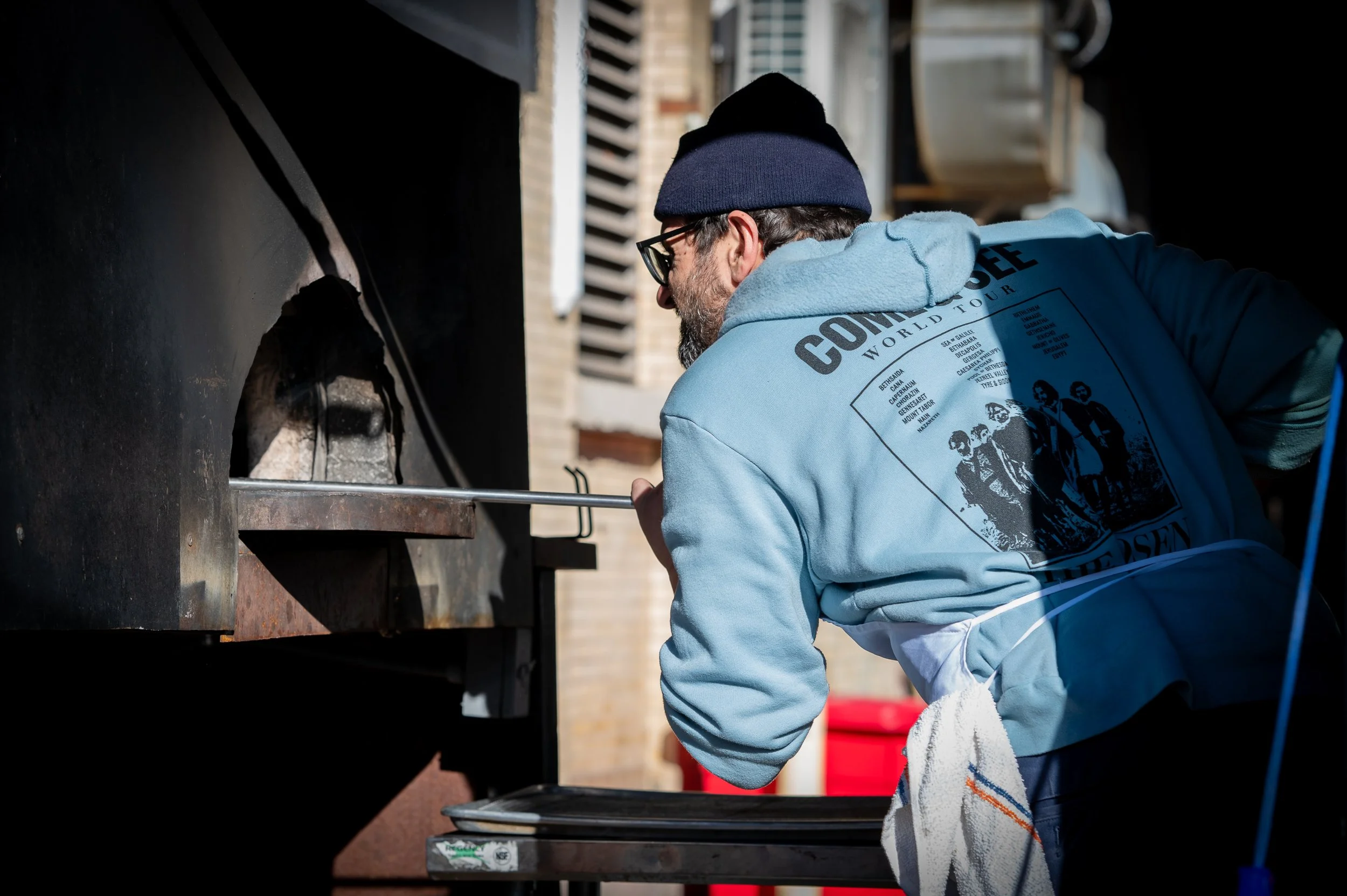 A man wearing a blue hoodie, glasses, and a navy beanie is cooking outdoors in front of a large black oven or smoker, using a metal poker to tend to the food inside.