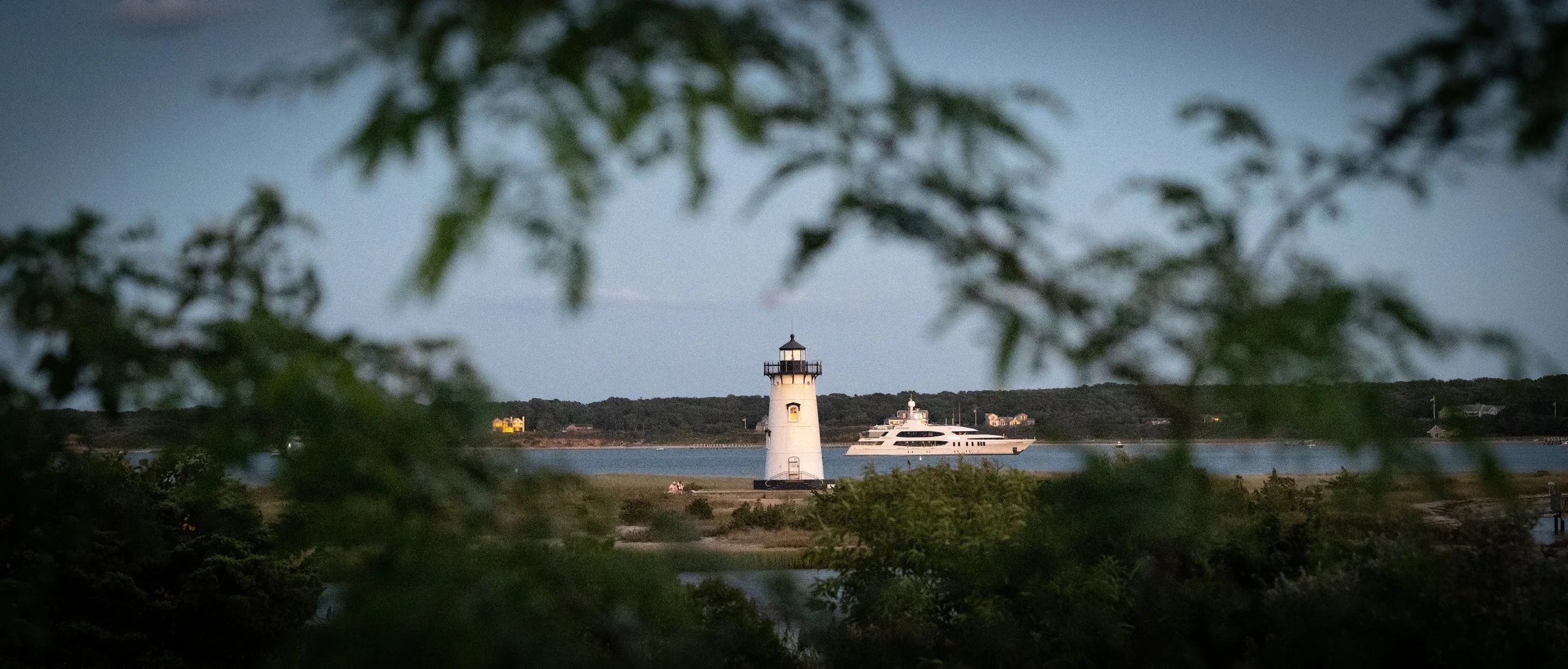 Lighthouse on a shoreline with a yacht passing by in the water, framed by blurred greenery in the foreground.