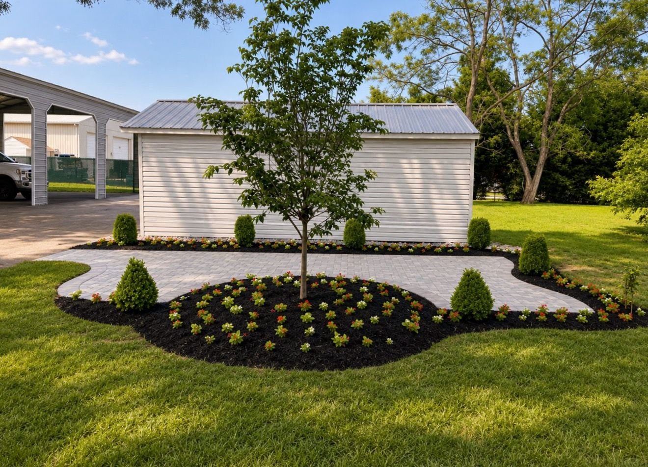Mulched planting beds with flowers and shrubs around a tree beside a lawn and white outbuilding