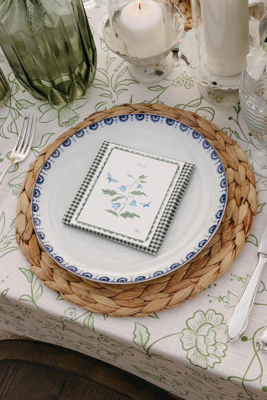 Charming place setting featuring a vintage blue dinner plate atop a woven charger, paired with a hand-painted menu card and a gingham-patterned napkin. The floral table linens and green glass accents add a refined garden-inspired touch to this elegan