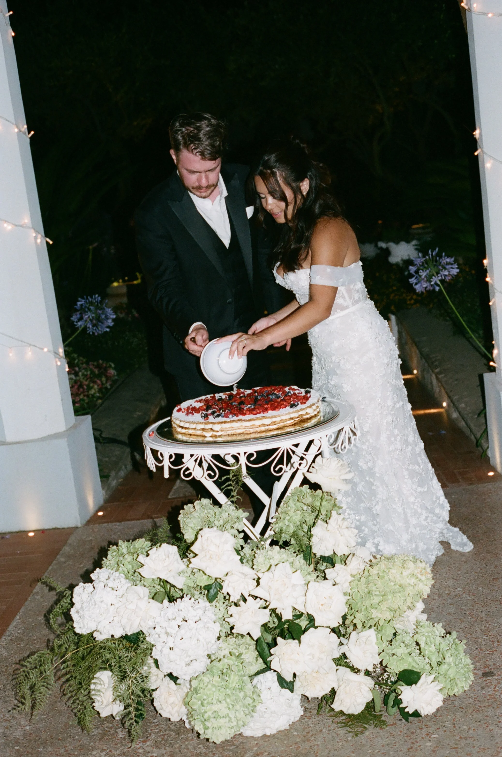 The bride and groom cutting their mille-feuille wedding cake, beautifully decorated with fresh fruit, at Villa Eva in Ravello, Italy. The cake is displayed on a vintage stand surrounded by lush floral arrangements, adding charm and elegance to this A
